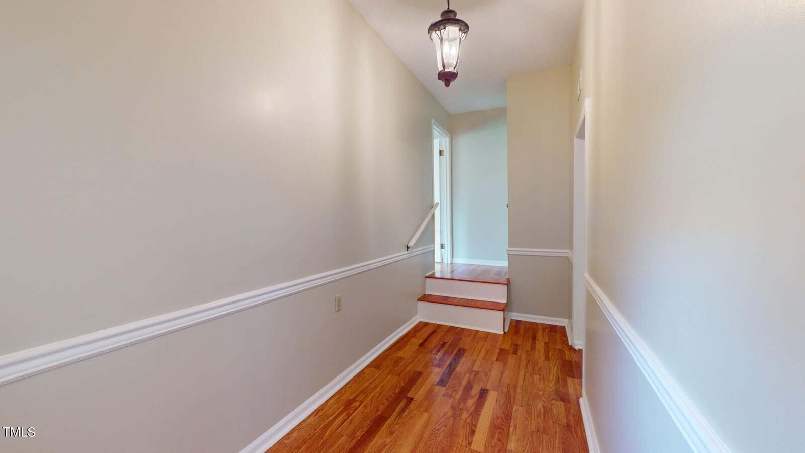 A hallway with light-colored walls, wooden flooring, white trim, a hanging light fixture, and a small set of stairs leading up to another room.