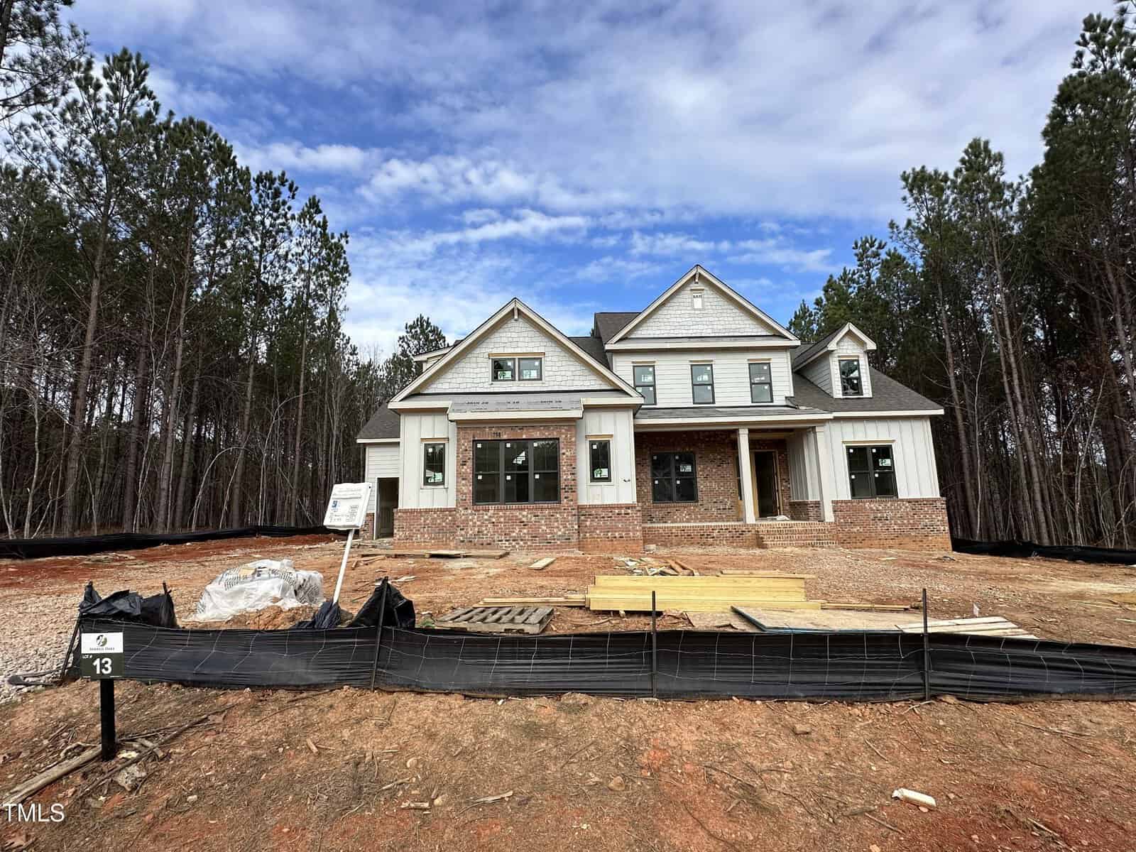 A two-story house under construction with a mix of brick and white siding exterior, surrounded by dirt, construction materials, and a black silt fence, with pine trees in the background.