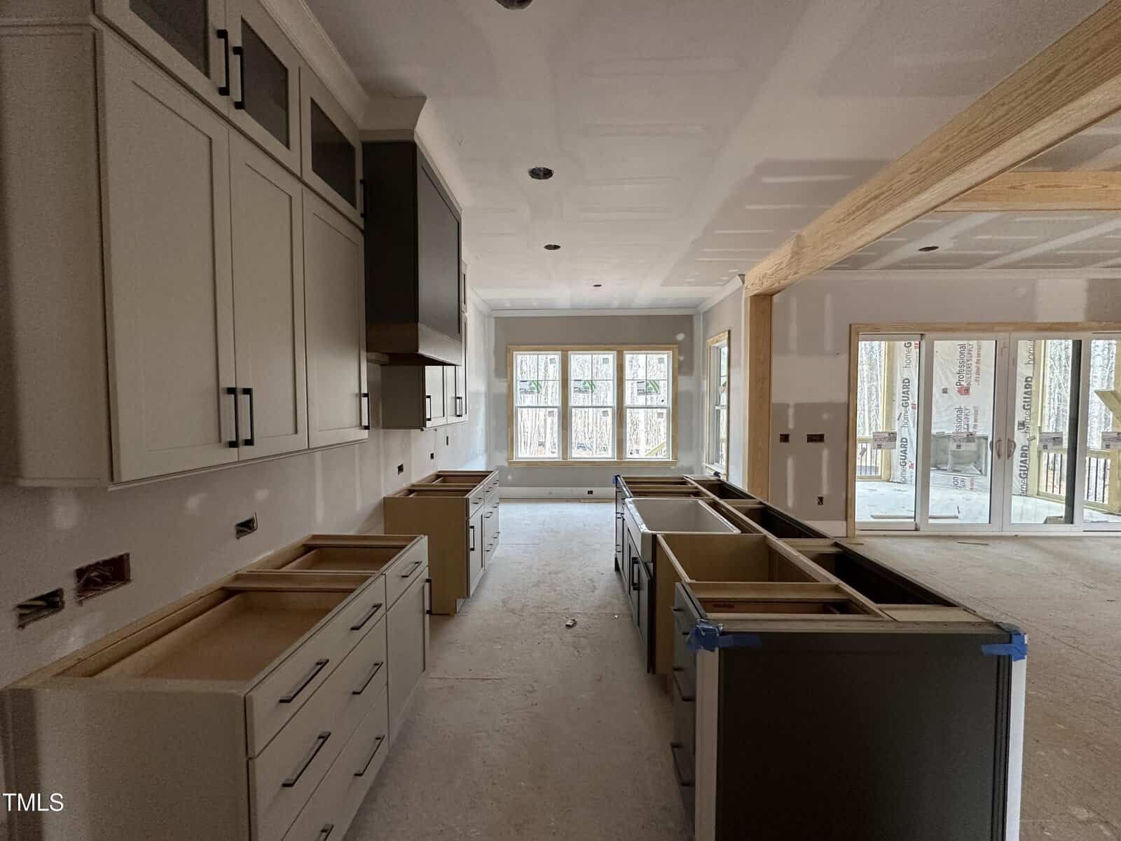 A kitchen under construction with unfinished cabinets and drawers installed, no countertops, and some appliances missing. The room is well-lit by large windows and there are exposed beams and unfinished walls.