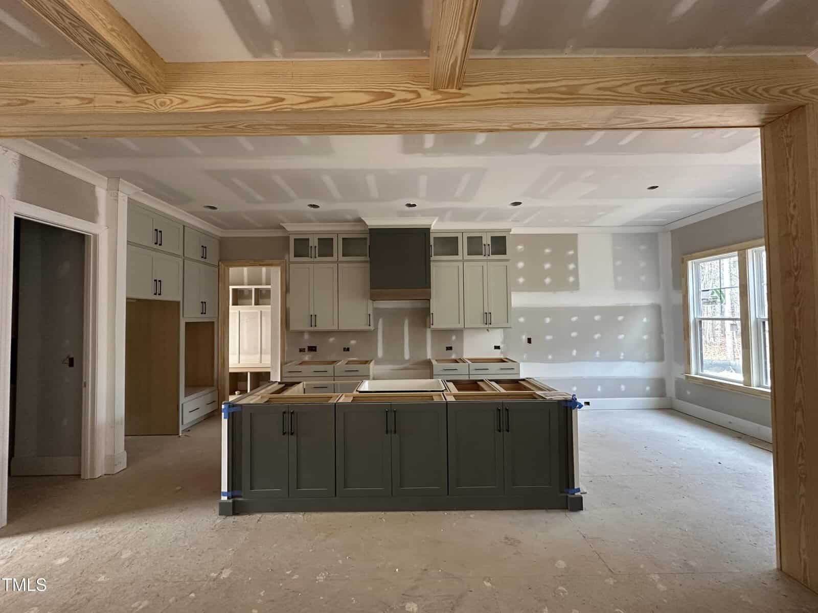 A kitchen under construction with unfinished floors, unpainted drywall, cabinets partially installed, a central island with dark green cabinetry, and large windows letting in natural light.