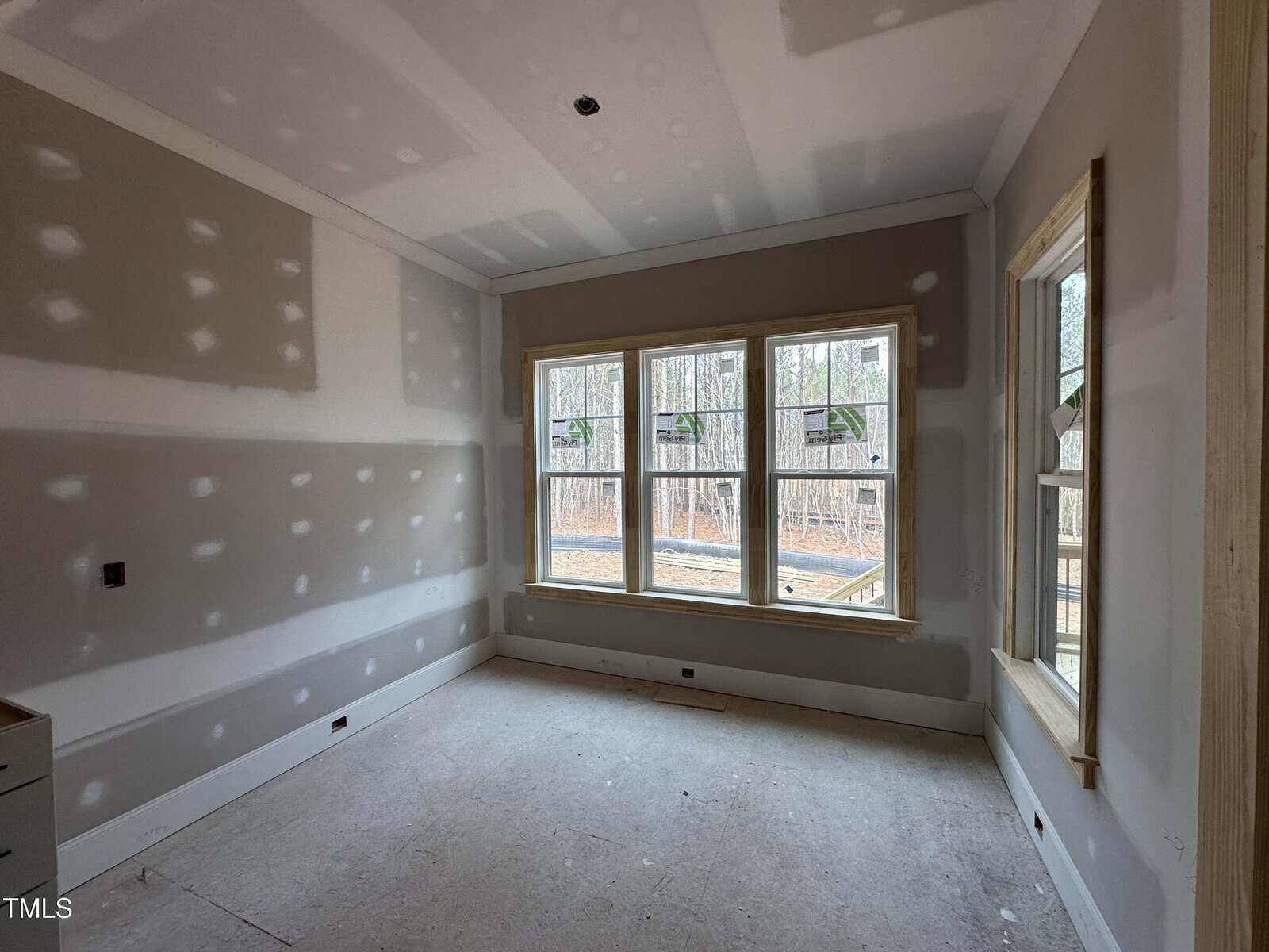 A room under construction with unfinished drywall on the walls and ceiling, large windows letting in natural light, and a view of trees outside. The floor is bare and there are no fixtures or furnishings.