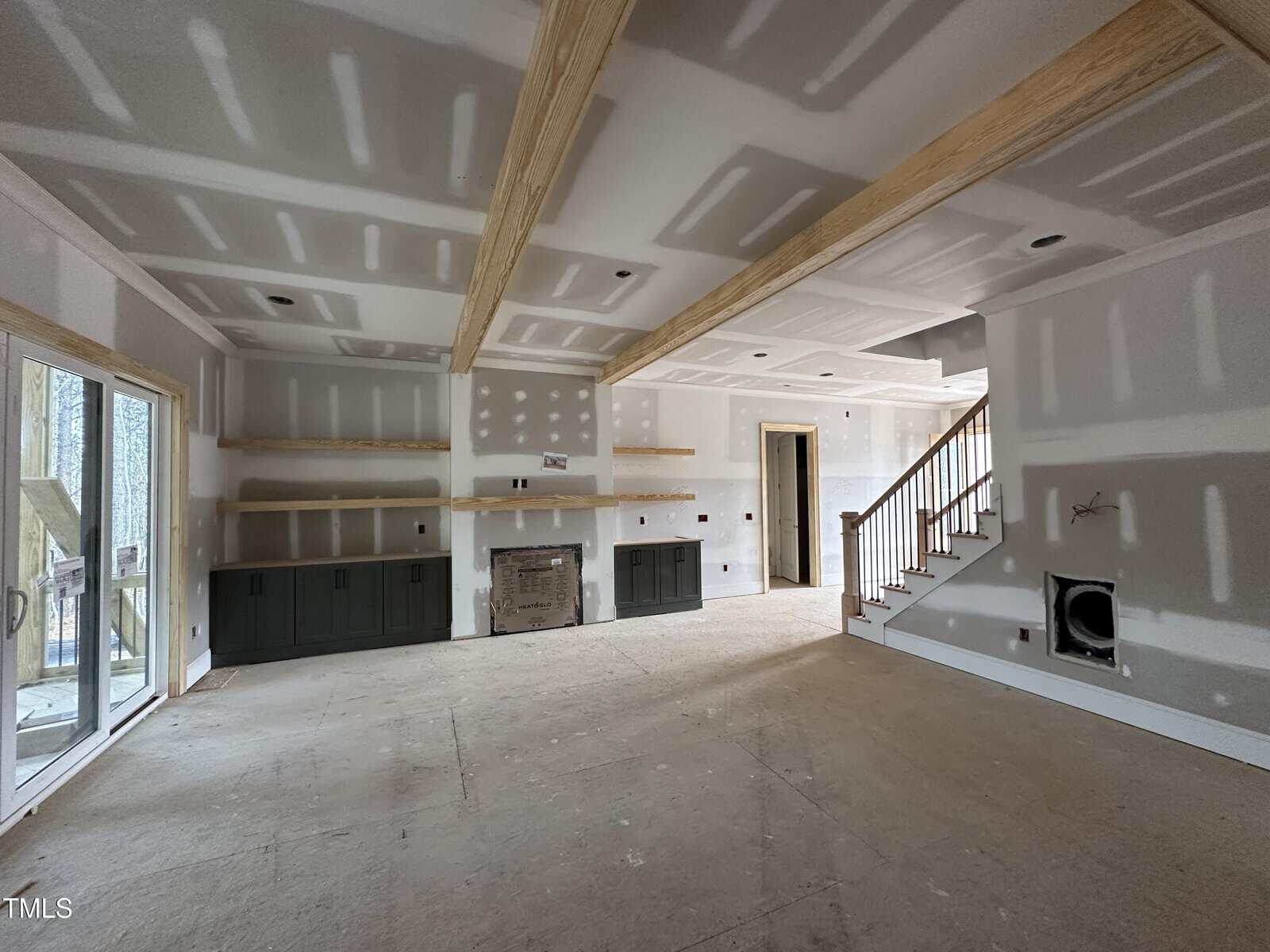Unfinished living room with drywall, exposed wooden ceiling beams, built-in shelves, and cabinets around a fireplace. A staircase is on the right, and a sliding glass door is on the left. No flooring or paint is completed.