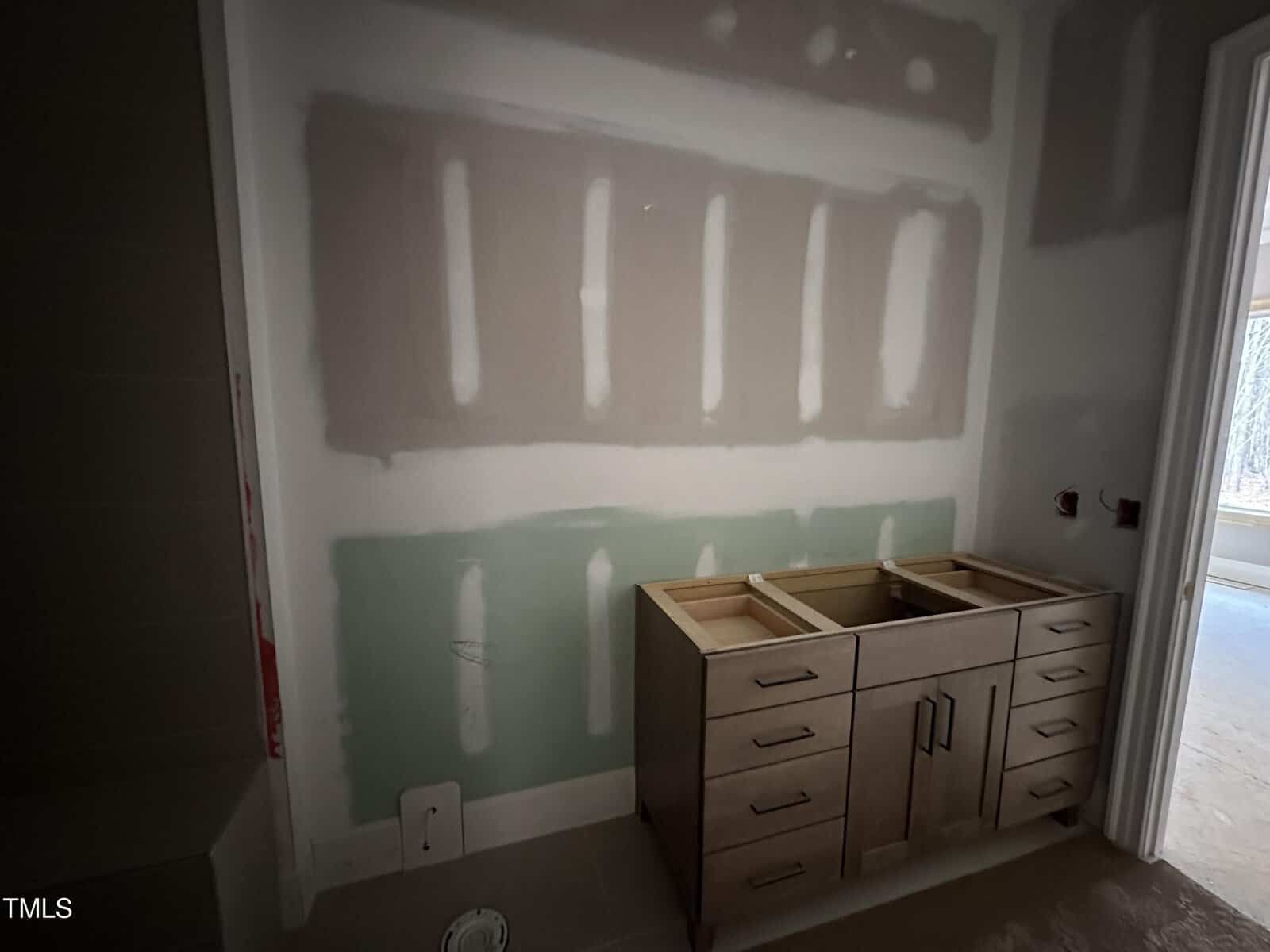 A bathroom under construction with unfinished drywall and a wooden vanity cabinet without a countertop, sink, or fixtures. Natural light enters from a window in another room.
