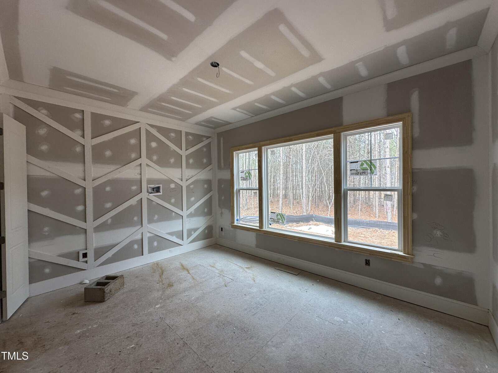 Unfinished room with drywall on walls and ceiling, large windows showing trees outside, and construction materials on the floor. Geometric pattern on one wall, no furniture present.