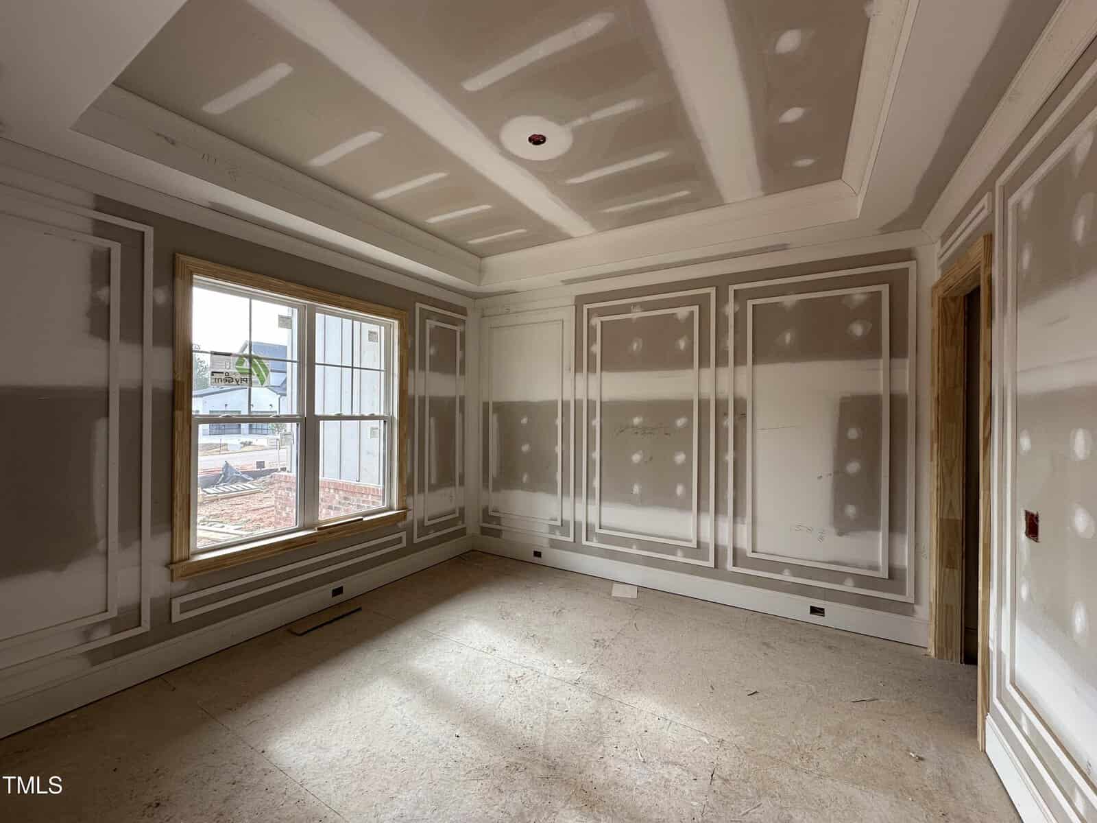 An unfinished room with drywall installed and seams taped, featuring a large window on one wall and intricate molding details on the walls and ceiling. The floor is bare subflooring.