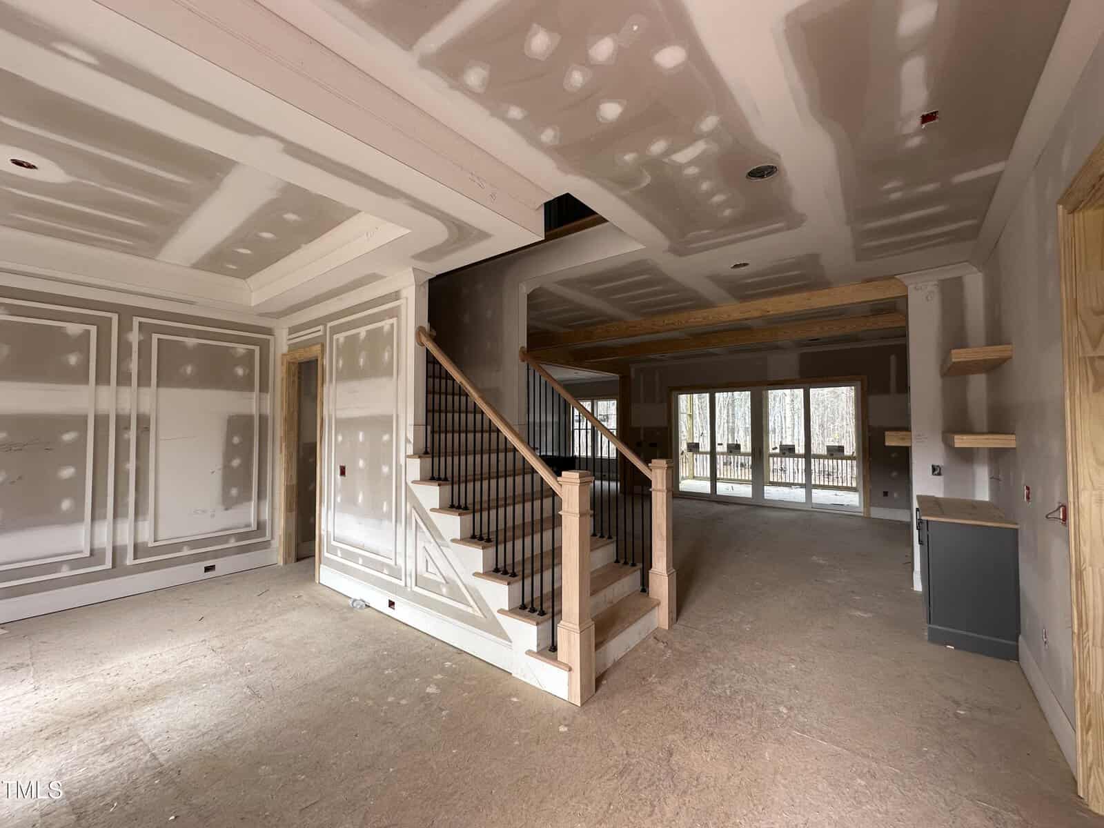 A large room in a house under construction with unfinished drywall, exposed wood stairs with black railings, unpainted doors, and large windows at the back letting in natural light.