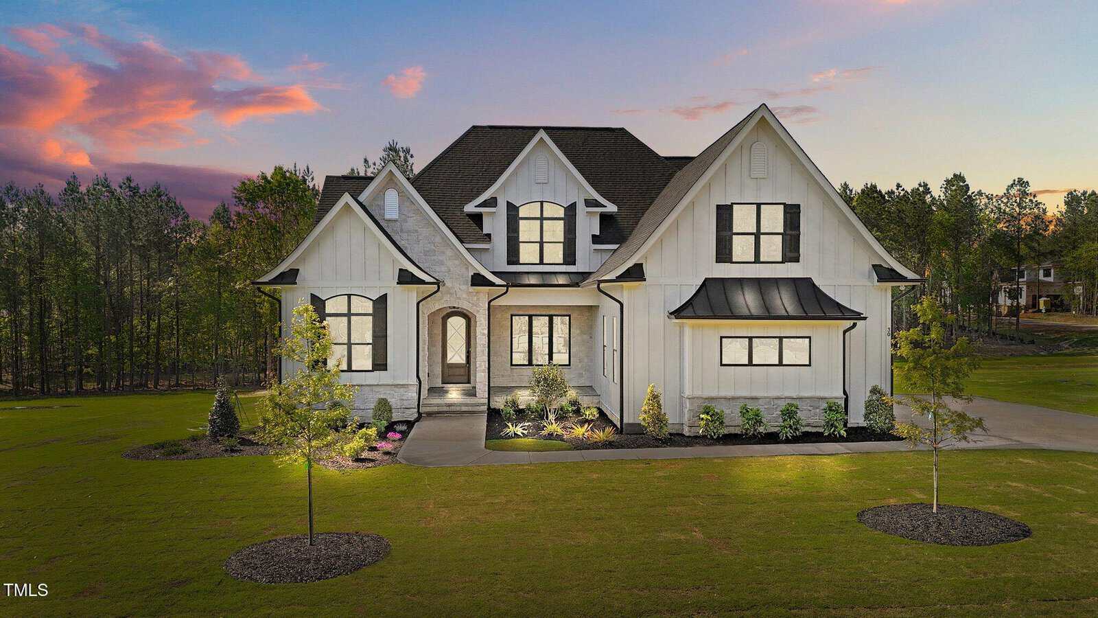 A two-story modern farmhouse with white siding, dark trim, and a gabled roof sits on a manicured lawn with young trees and landscaped shrubs, surrounded by tall trees at sunset.