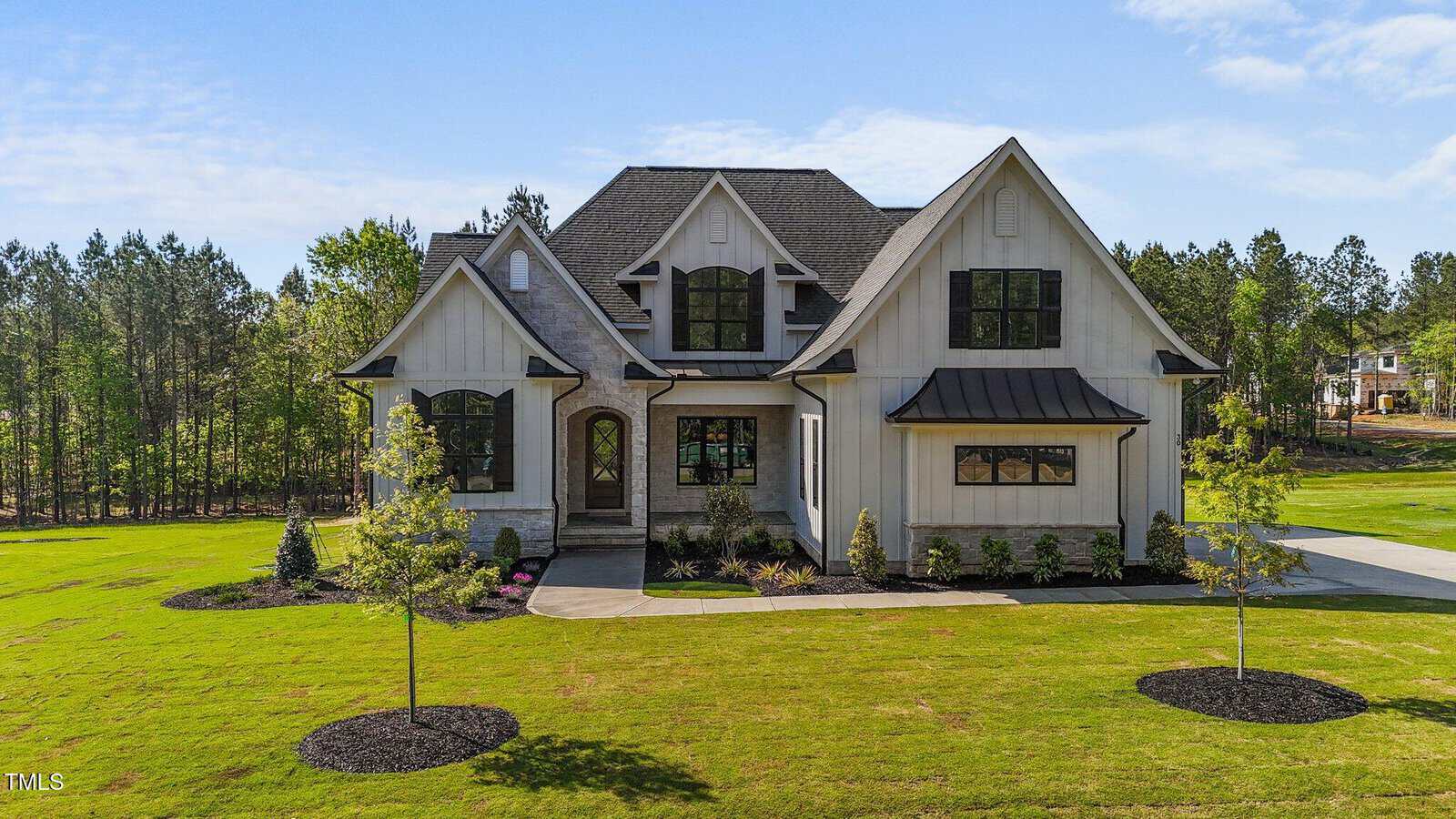 A two-story modern farmhouse with light-colored siding, dark trim, multiple gables, and a manicured front lawn with young trees and shrubs. Woods and other homes are visible in the background.