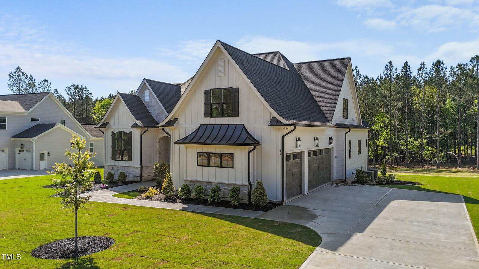 A modern two-story white house with a black roof, two-car garage, manicured lawn, landscaped plants, and a concrete driveway, situated in a suburban neighborhood with trees in the background.