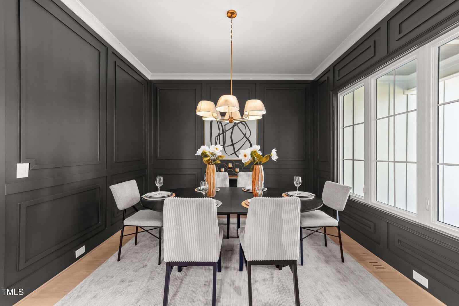A modern dining room with dark paneled walls, a round table set for four, light upholstered chairs, a central gold chandelier, and large windows letting in natural light. White flowers and tableware decorate the table.