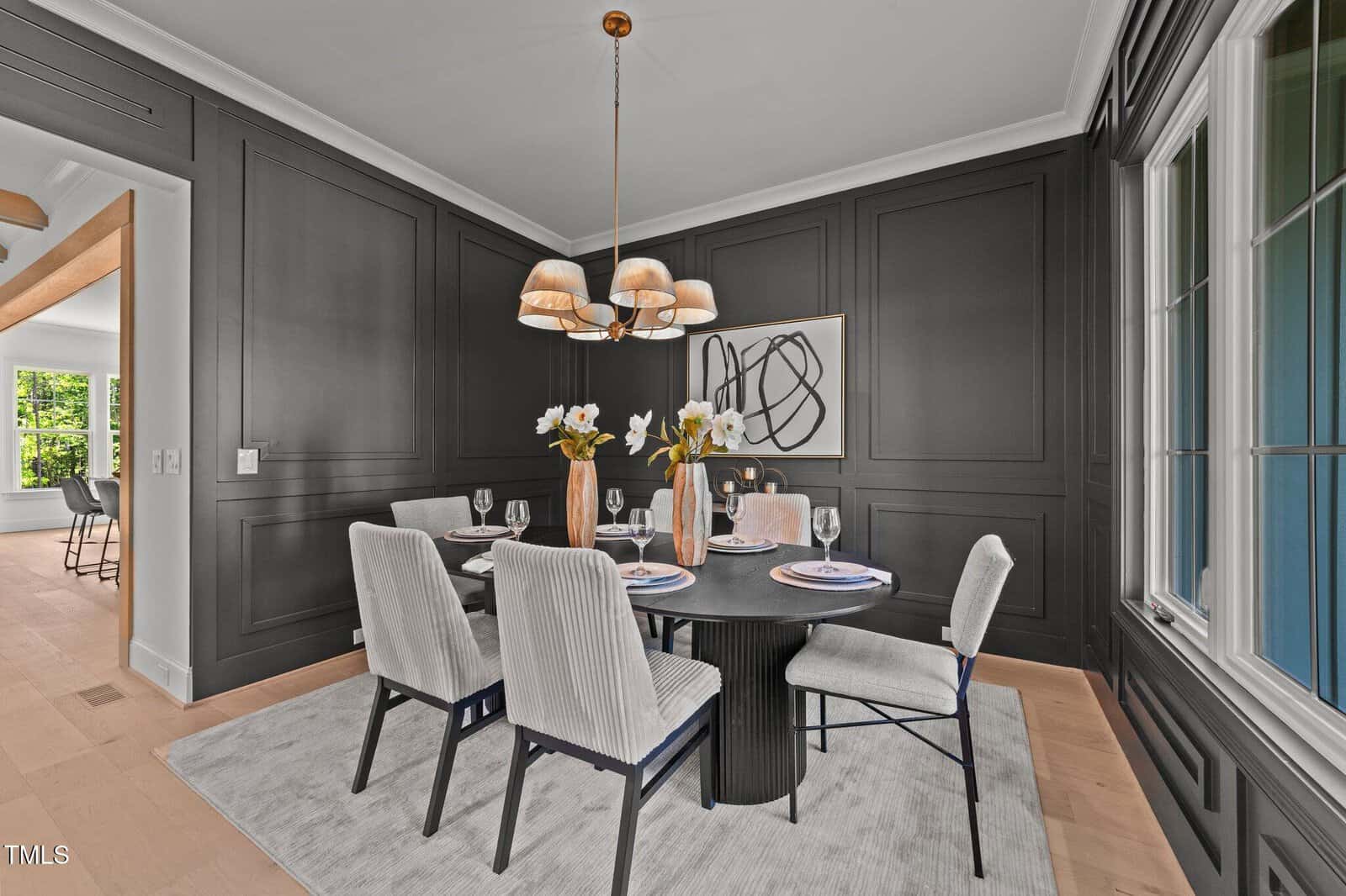 A modern dining room with dark paneled walls, a round black table set for six, light upholstered chairs, a beige rug, and a pendant light fixture above. A large abstract artwork hangs on the wall.