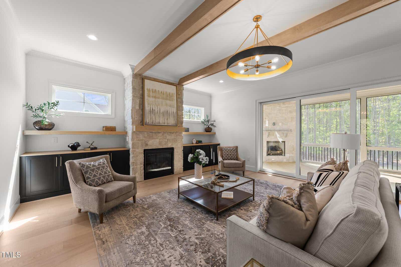 Modern living room with exposed wood beams, stone fireplace, built-in shelves, armchairs, a beige sofa, coffee table, and large windows showing a patio and trees outside. Neutral decor and ample natural light.