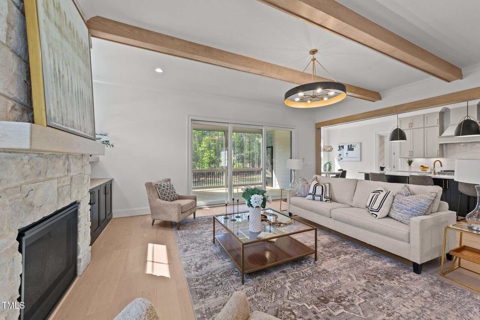 Modern living room with exposed wood beams, a stone fireplace, neutral-toned furniture, a patterned rug, sliding glass doors to a deck, and an open view into a white kitchen with barstools and pendant lights.