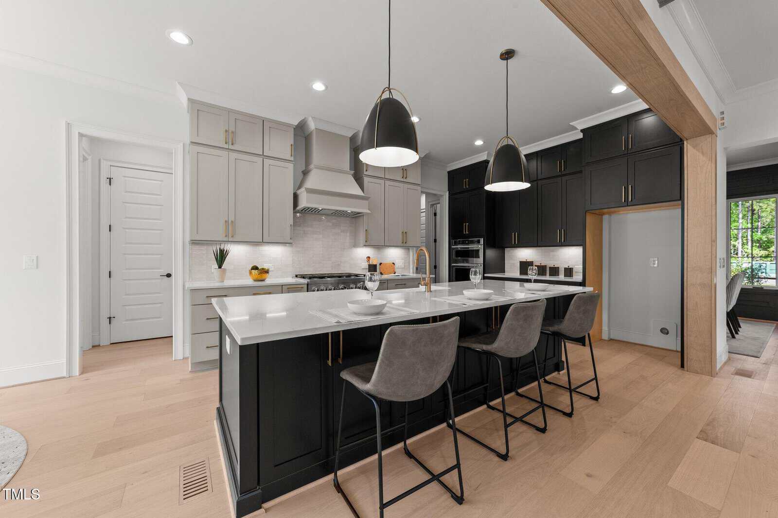 Modern kitchen with light wood floors, a large white island with three gray barstools, gray and black cabinets, stainless steel appliances, two pendant lights, and a view of a window with greenery outside.