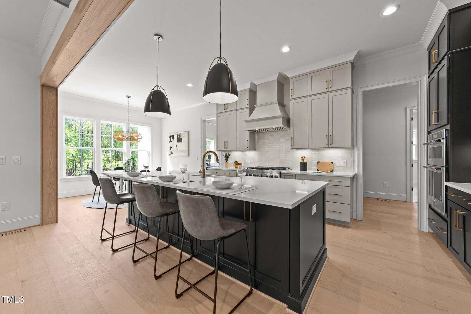 Modern kitchen with light wood floors, a large island with black base and white countertop, three gray barstools, pendant lights, stainless steel appliances, and light gray cabinets. Large windows provide natural light.