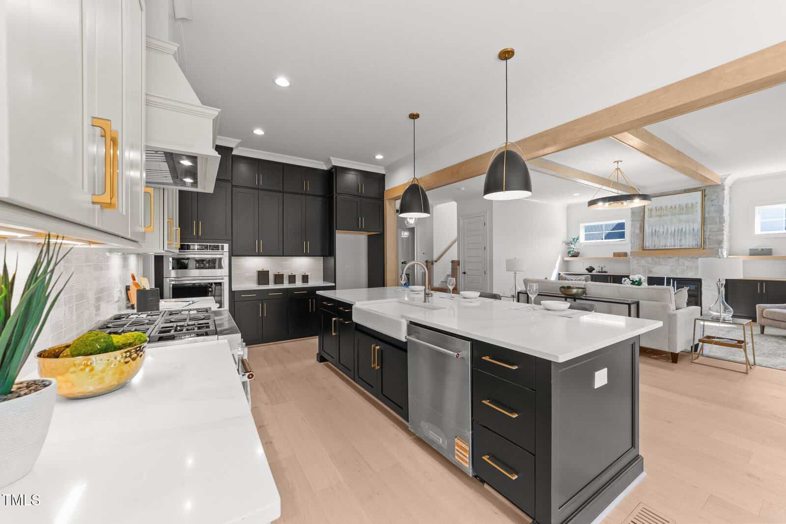 Modern kitchen with black cabinets, white countertops, built-in stainless steel appliances, a large island with sink, pendant lights, and open layout leading to a bright living and dining area with light wood flooring.
