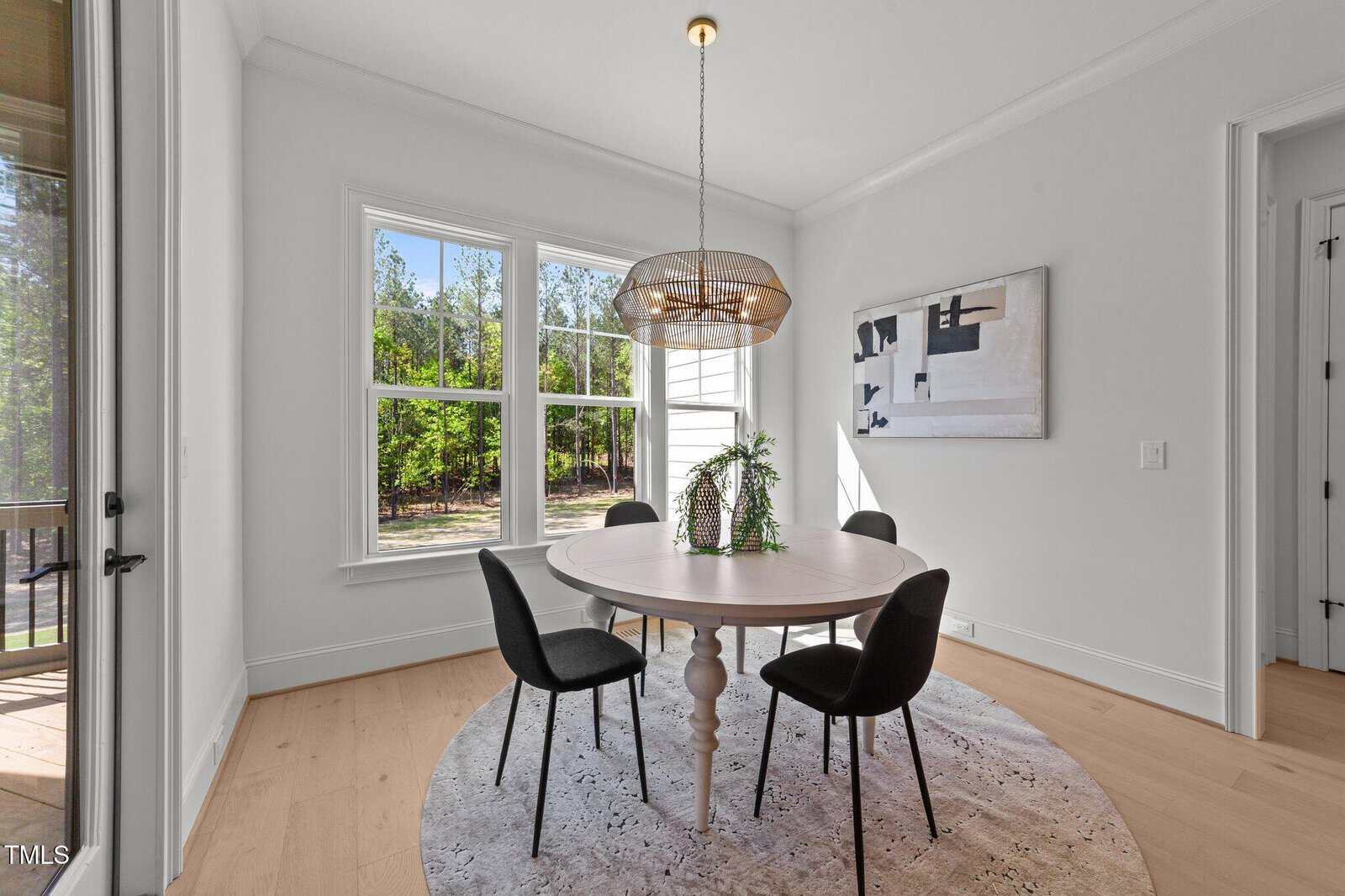 A round dining table with four black chairs sits on a circular rug in a bright room with large windows, a modern light fixture, and abstract wall art. Trees and greenery are visible outside the window.