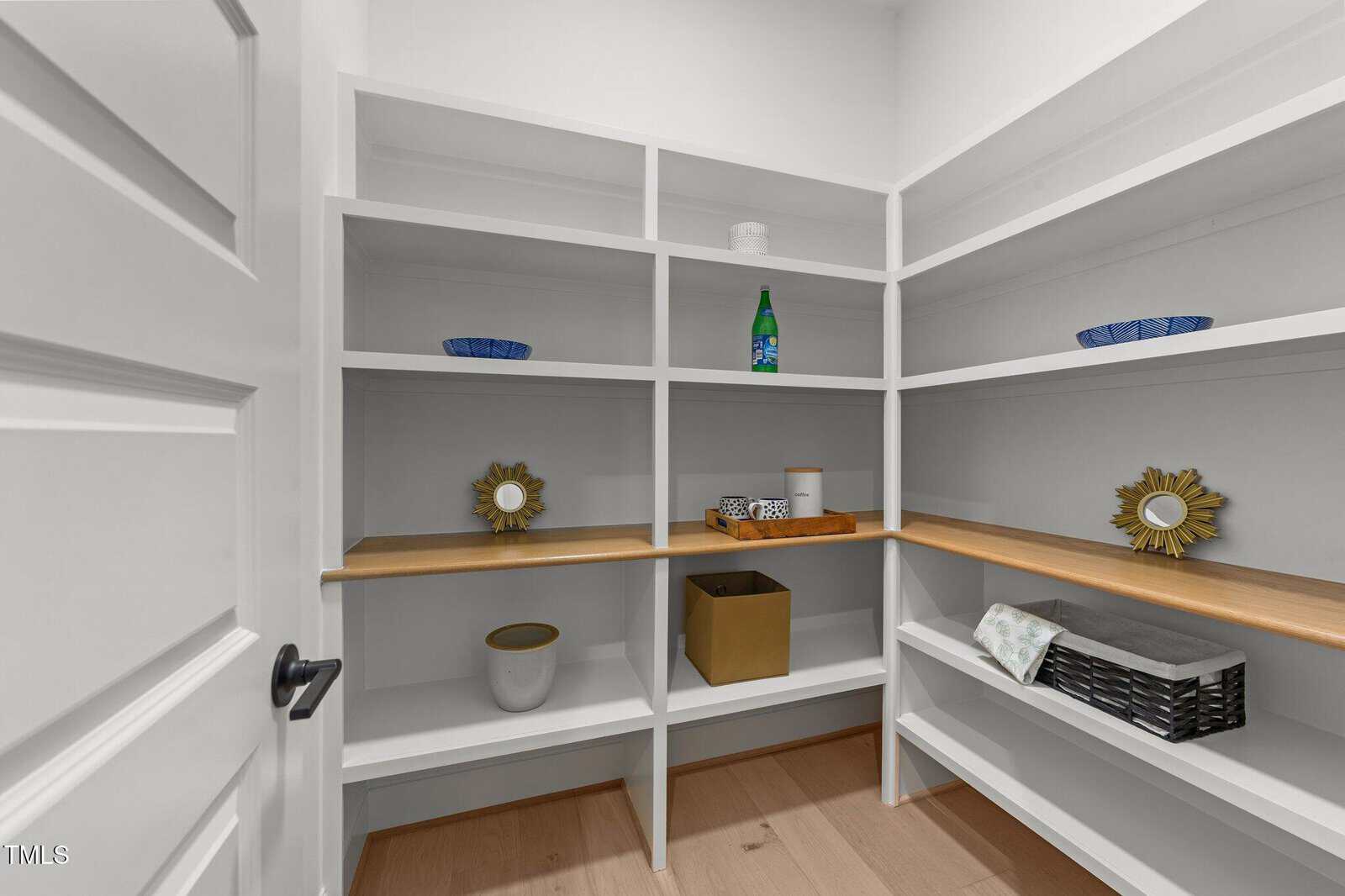 A walk-in pantry with white built-in shelves, some decorated with baskets, bowls, a green bottle, mirrors, a towel, and containers. The shelves are mostly empty and the floor is light wood.