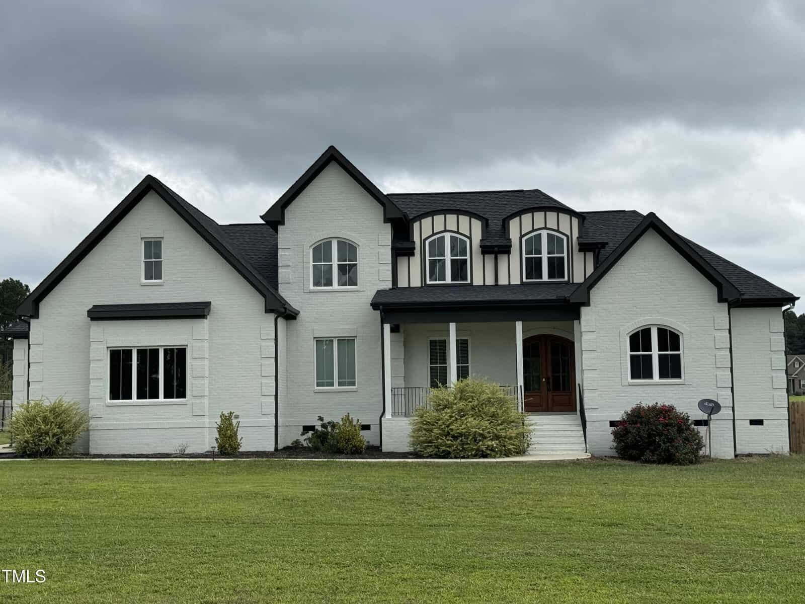 A large, two-story white brick house with black trim, multiple gables, arched windows, and a covered front porch, set on a spacious green lawn under a cloudy sky.