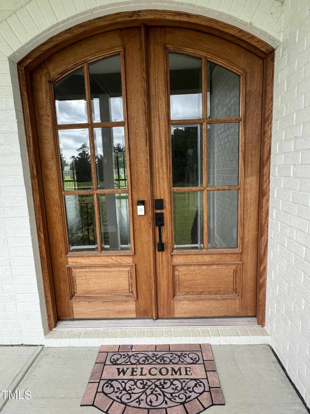 A wooden double front door with glass panes and black hardware is set in a white brick wall. A decorative "WELCOME" doormat is placed in front of the door on a light-colored porch.