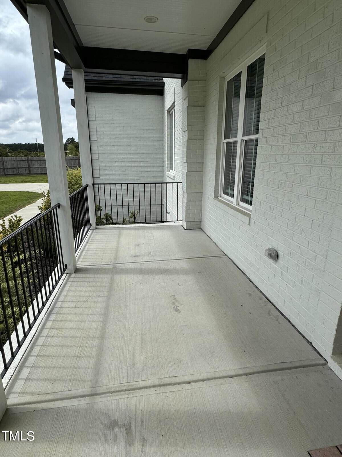A covered front porch with light-colored brick walls, black railing, large window, and concrete floor. The porch overlooks a green lawn and is partially shaded by the roof.