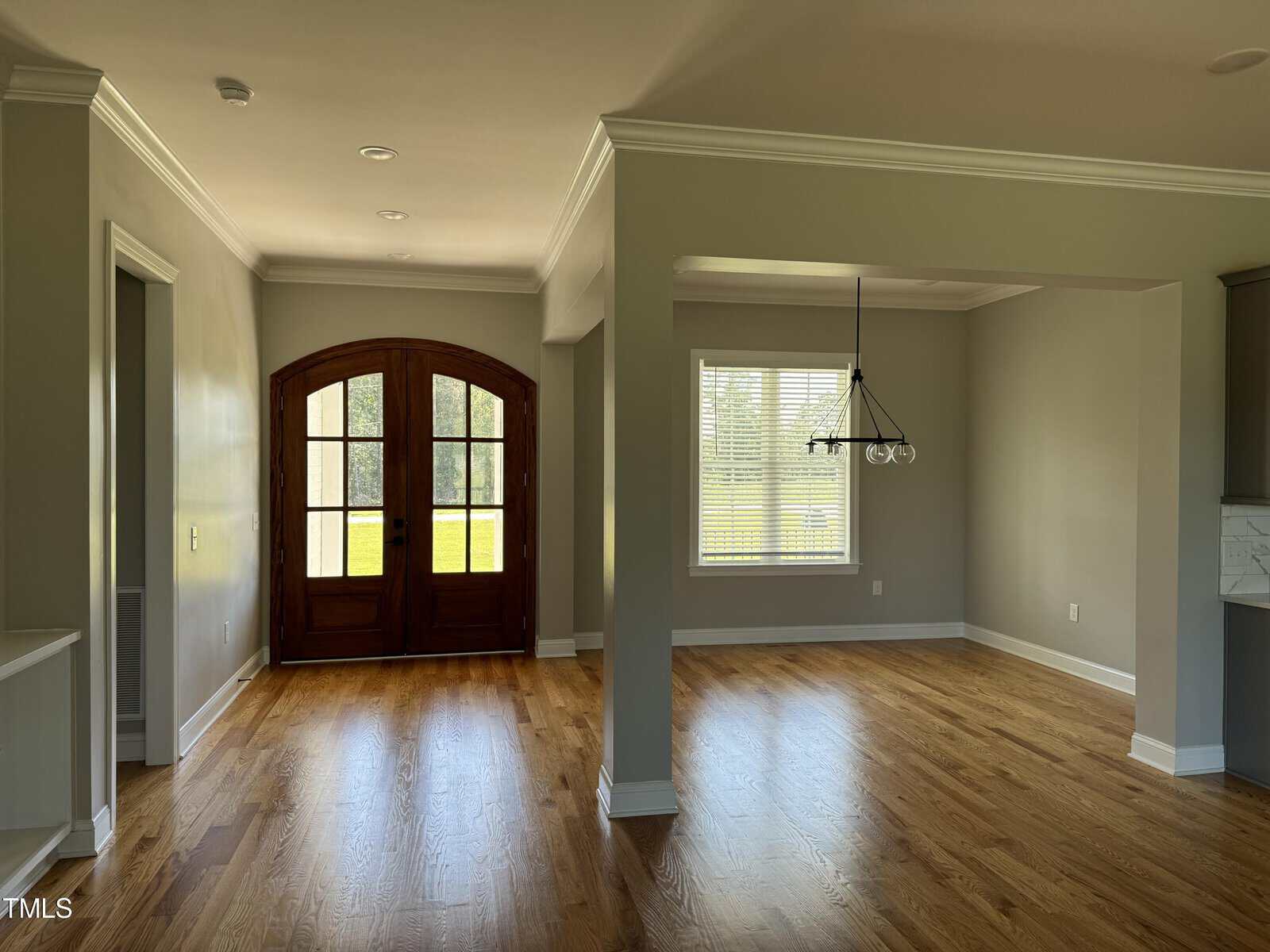 A bright entryway with wooden double doors, large windows, and an adjacent dining area with an overhead light fixture. The space features light gray walls, white trim, and hardwood floors.