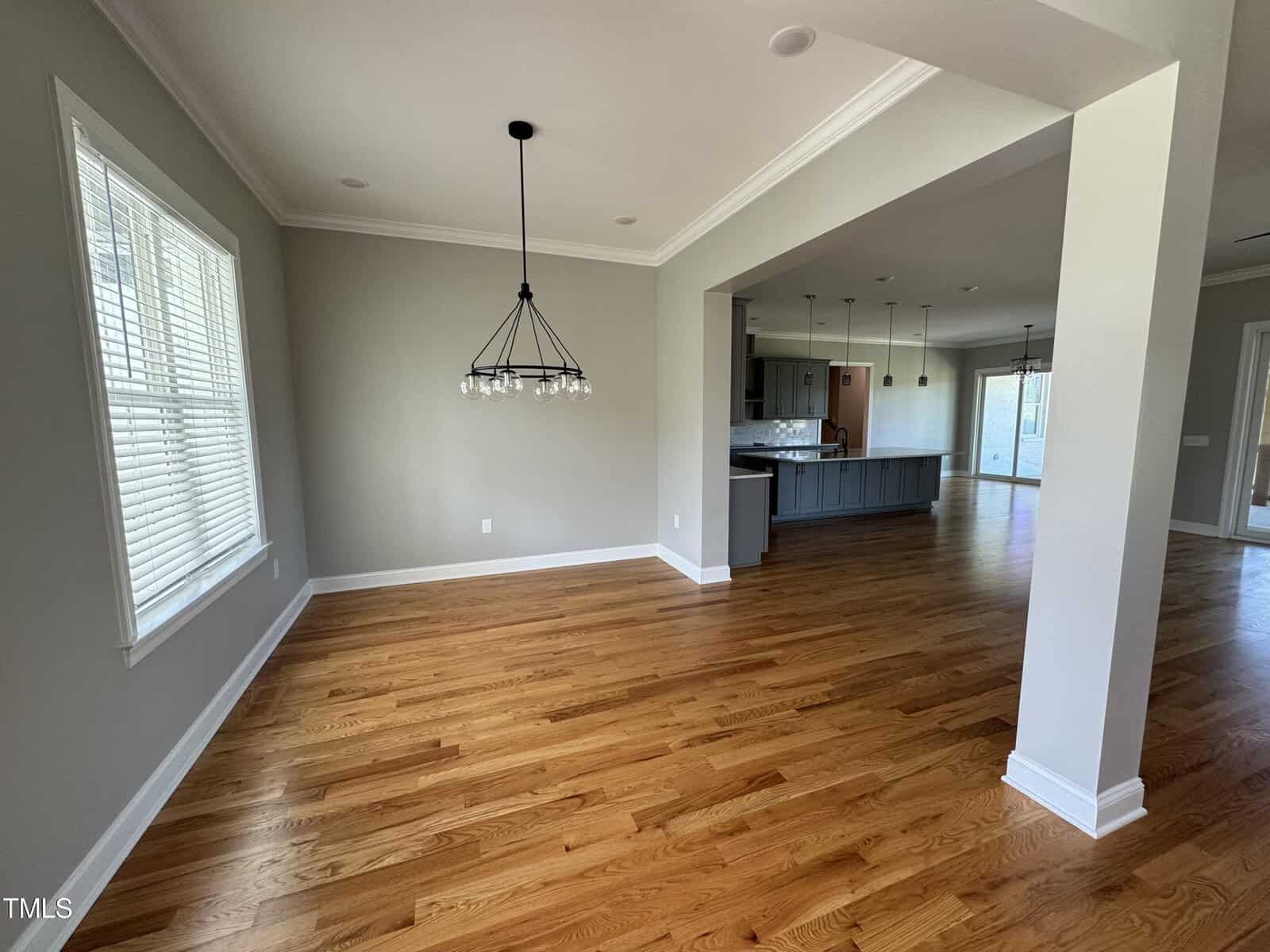 Spacious open-concept room with hardwood floors, neutral walls, large windows, a modern chandelier, and a view into a kitchen with pendant lights and a central island.
