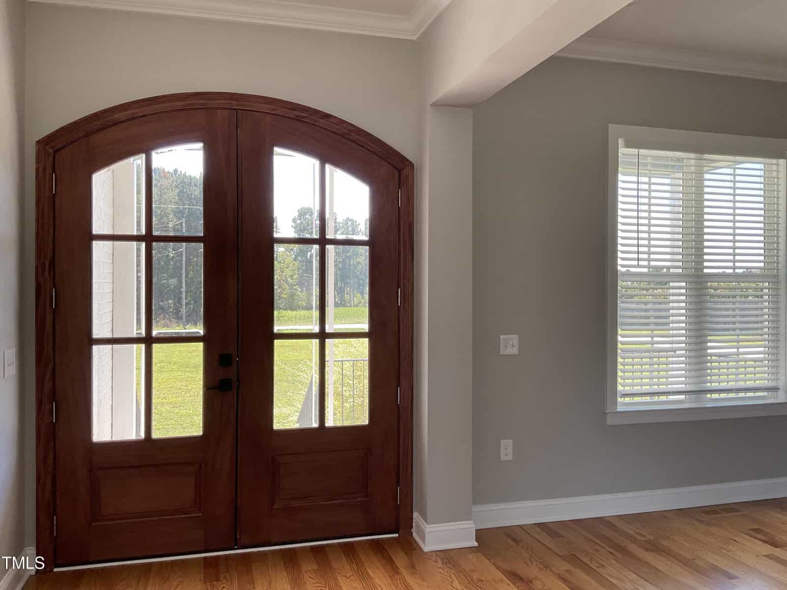 A bright entryway with double wooden arched doors featuring glass panels, adjacent to a room with a large window with white blinds, light gray walls, and hardwood floors. Sunlight streams in from outside.