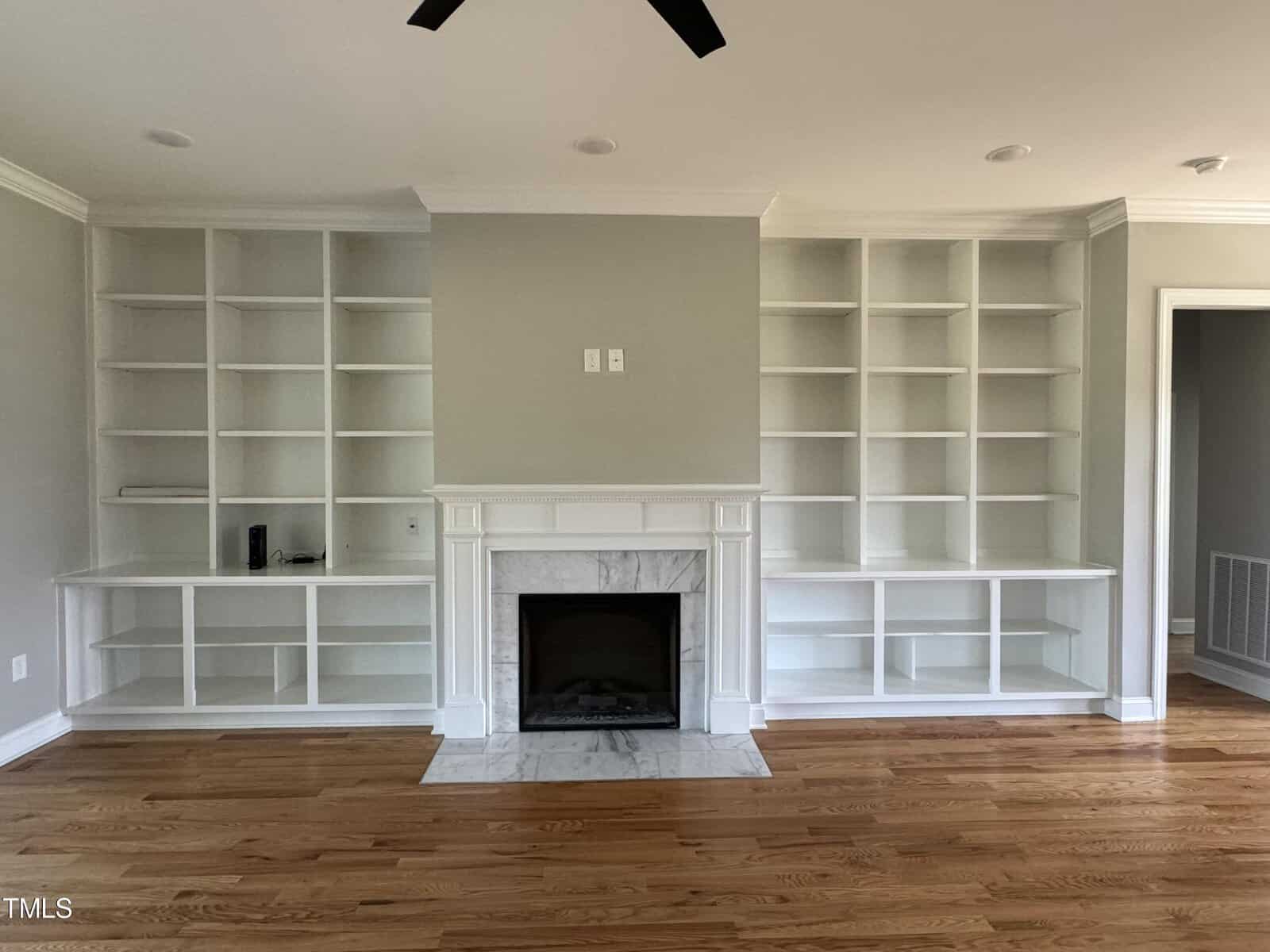 A living room with built-in white bookshelves on either side of a white fireplace with a marble surround, set against light gray walls and hardwood floors. The shelves are empty.