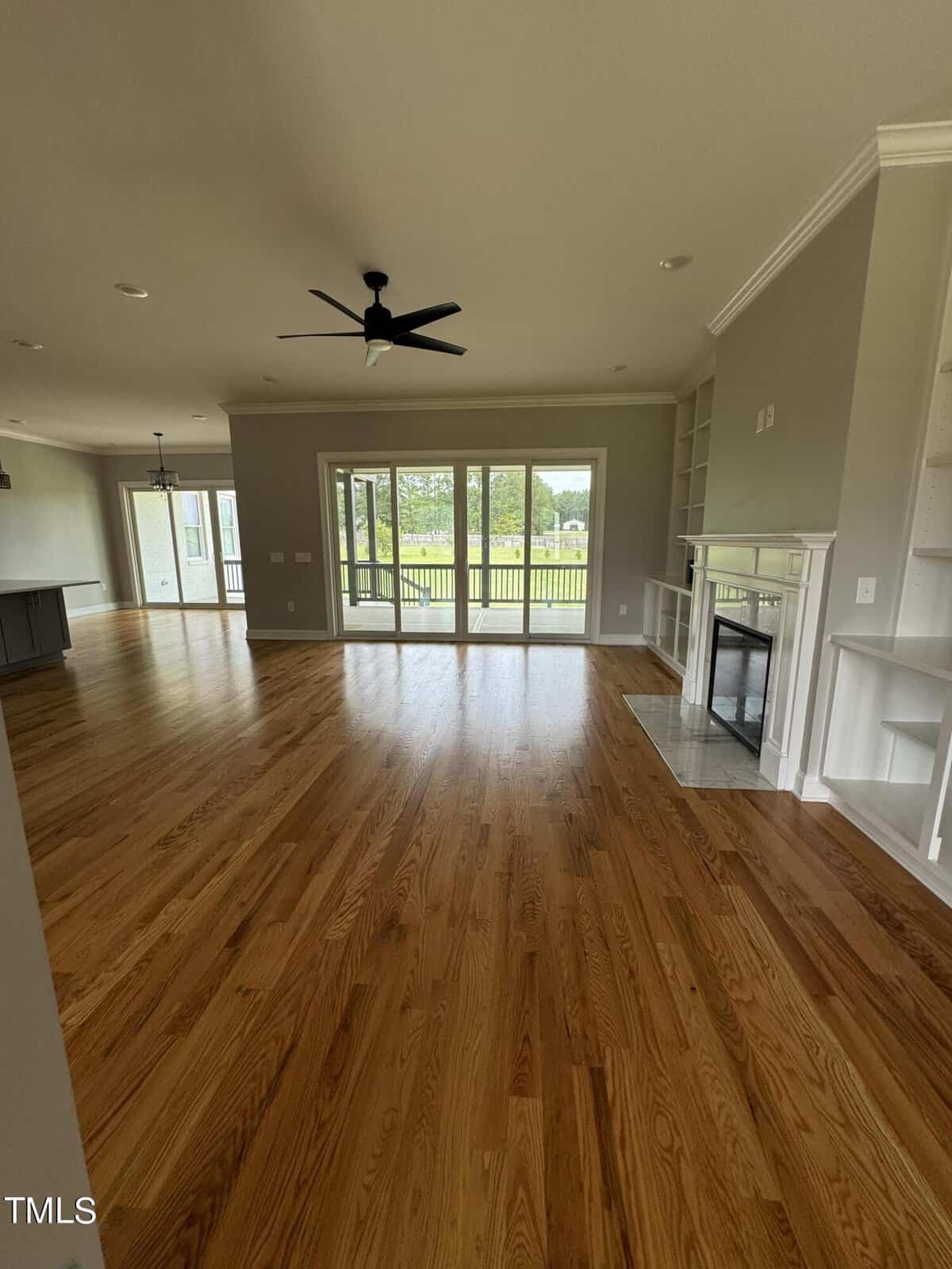 Spacious living room with hardwood floors, a ceiling fan, built-in shelves, a fireplace, and large glass doors opening to a patio with a view of a green lawn and trees.