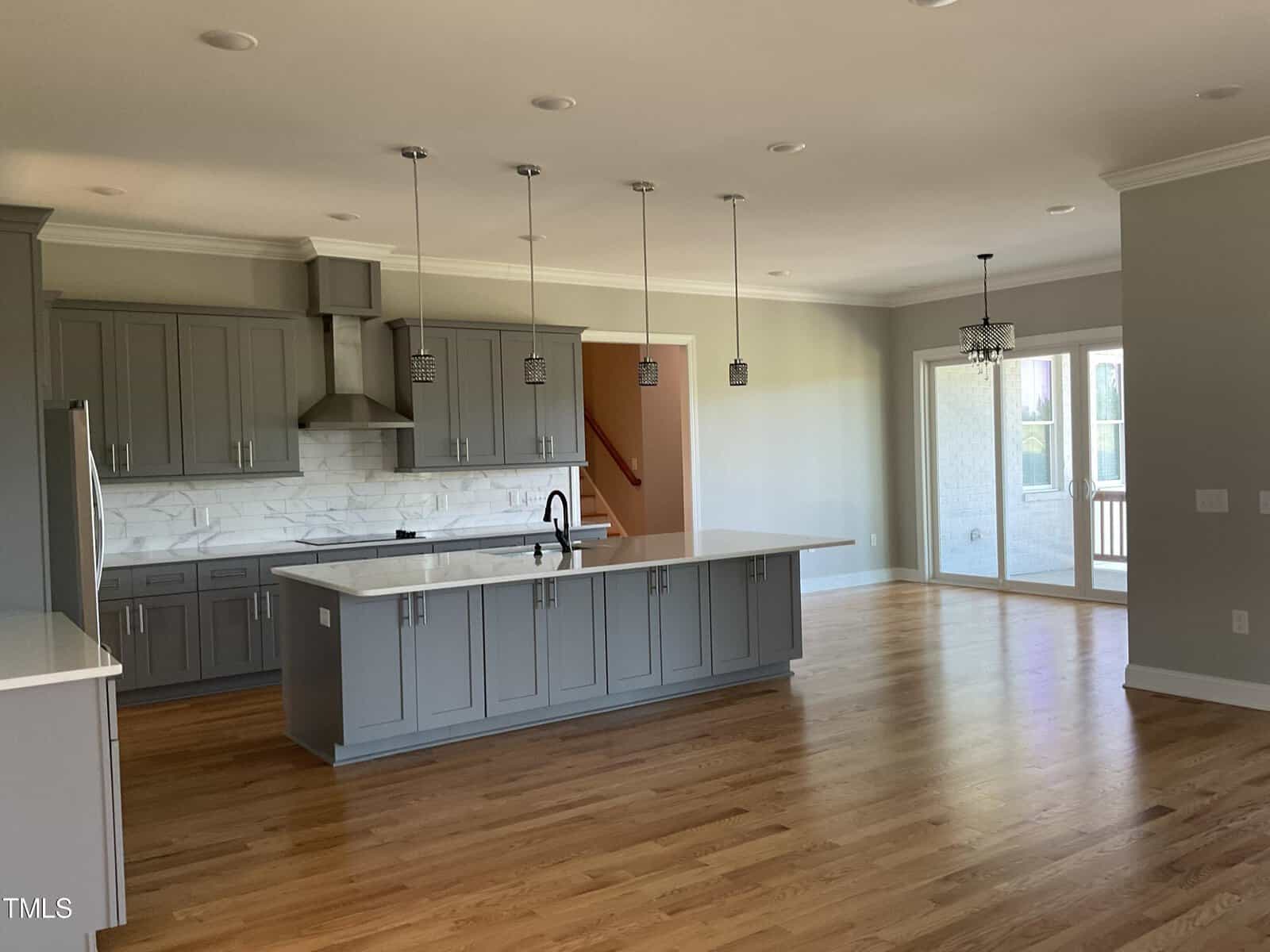 A modern kitchen with gray cabinets, a white marble backsplash, a large island with a sink, stainless steel appliances, and pendant lights, next to an open dining area with hardwood floors and sliding glass doors.