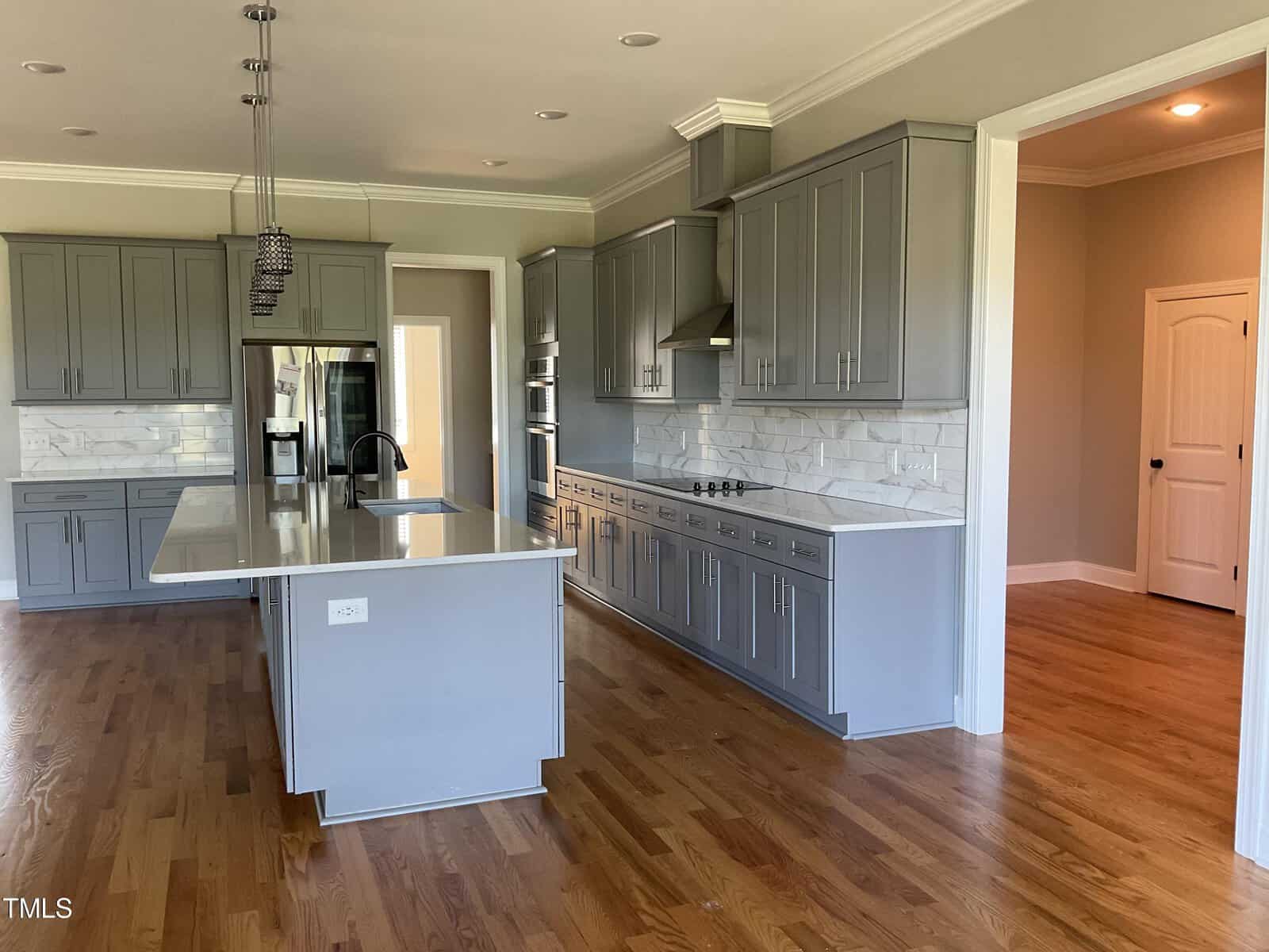 Modern kitchen with gray cabinets, white countertops, tile backsplash, stainless steel appliances, island with sink, pendant lighting, and hardwood floors. Doorways and natural light are visible in the background.