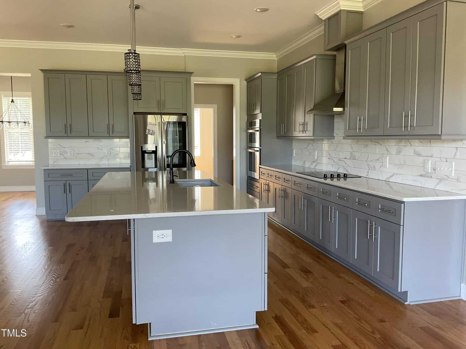 Modern kitchen with gray cabinets, white countertops, island with sink, stainless steel appliances, stove, and light wood floors. Marble-style backsplash and pendant lights add a contemporary touch.