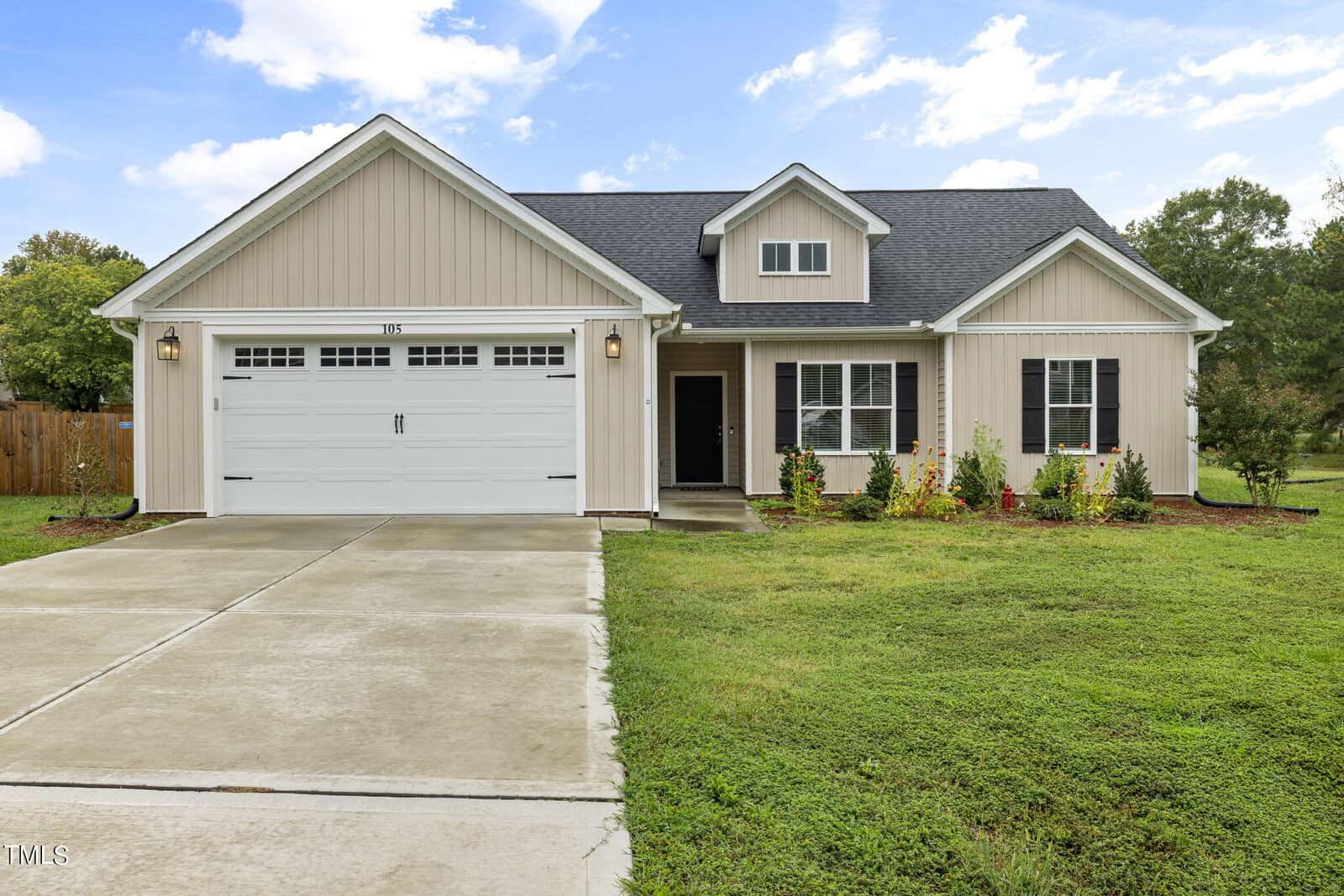A beige single-story house with a dark gray roof, two-car garage, and a well-kept front yard. The driveway leads to a wide garage door with small windows. There are bushes and a small lawn in front, under a partly cloudy sky.