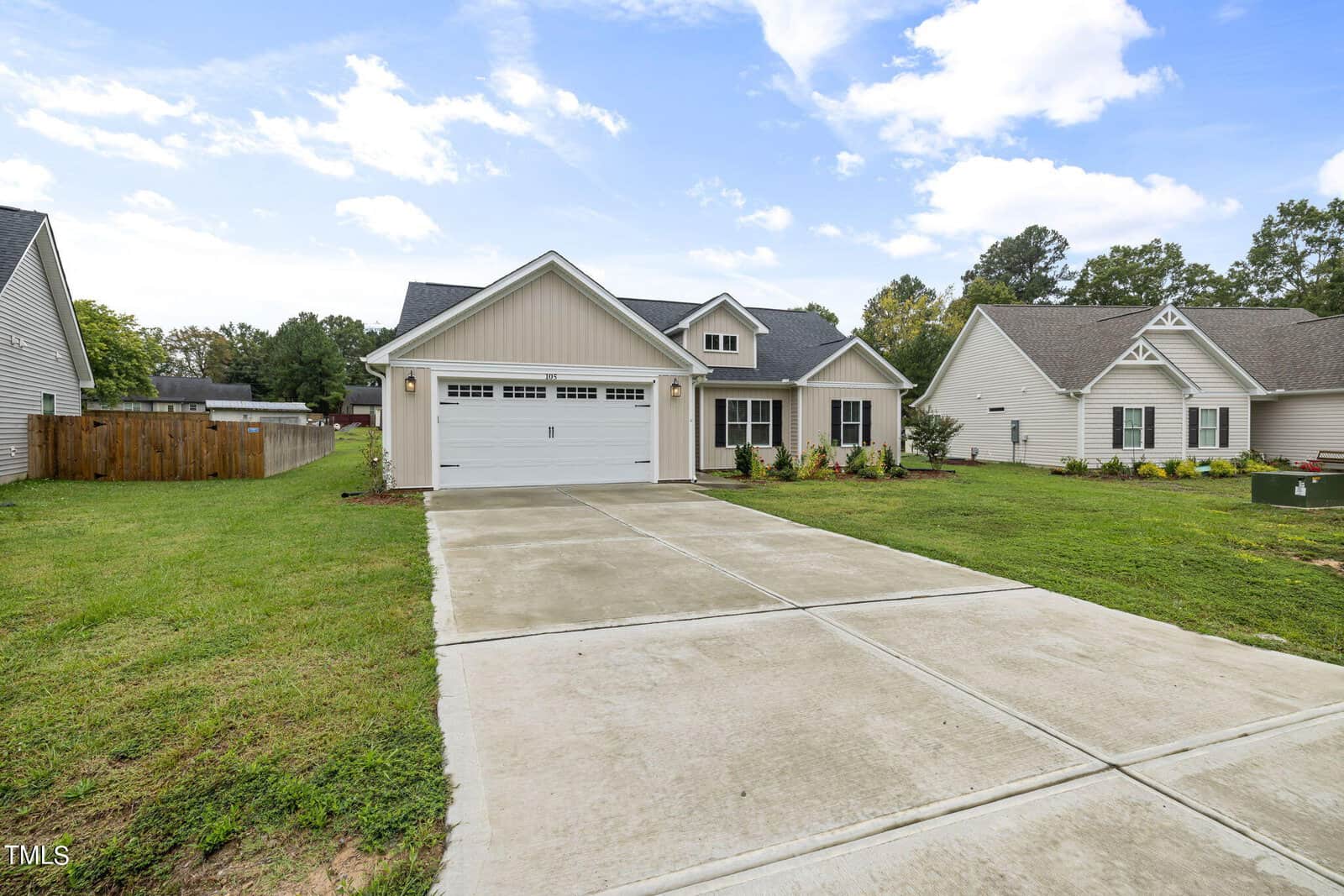 A single-story suburban house with a gray exterior and white trim. The house has a two-car garage and a concrete driveway. The front yard is grassy, and other similar houses are visible on either side. The sky is partly cloudy.