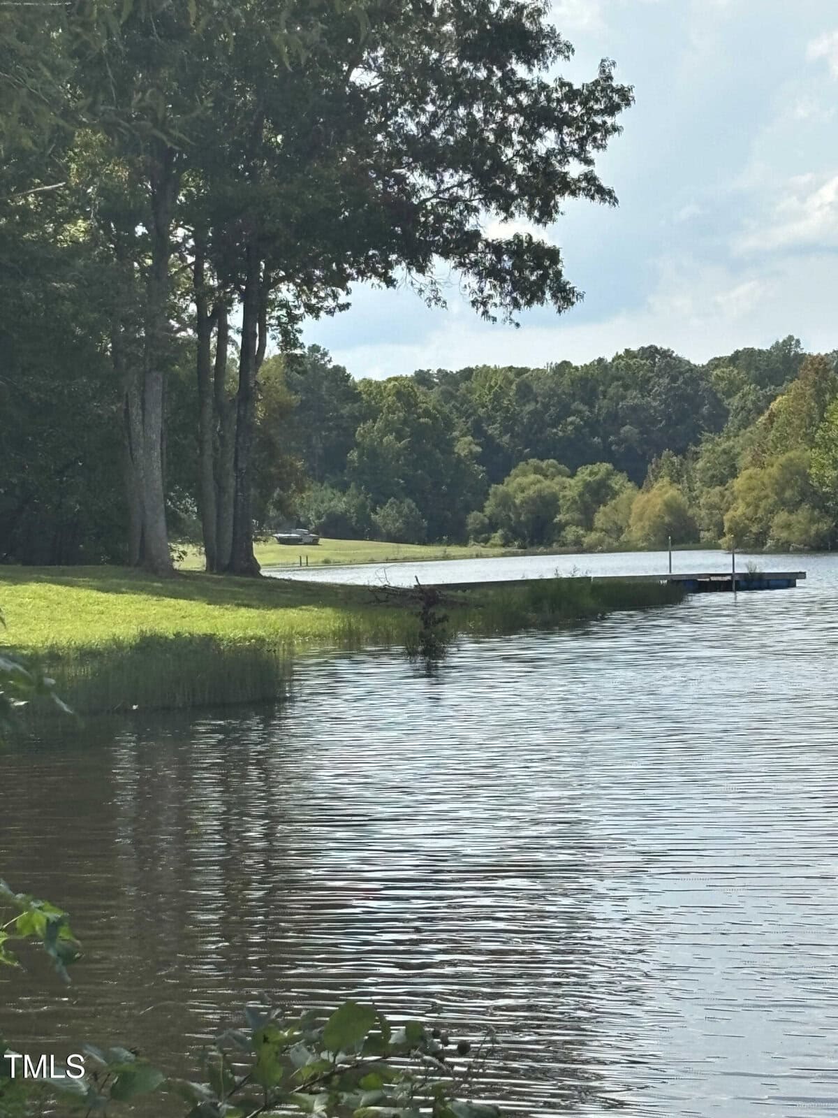 A tranquil lake scene along 9535 Catfish Trail, with trees lining the shoreline on a sunny day. A small dock extends over the water, and the sky is partly cloudy. The landscape is beautifully reflected in the rippling water near Oxford.