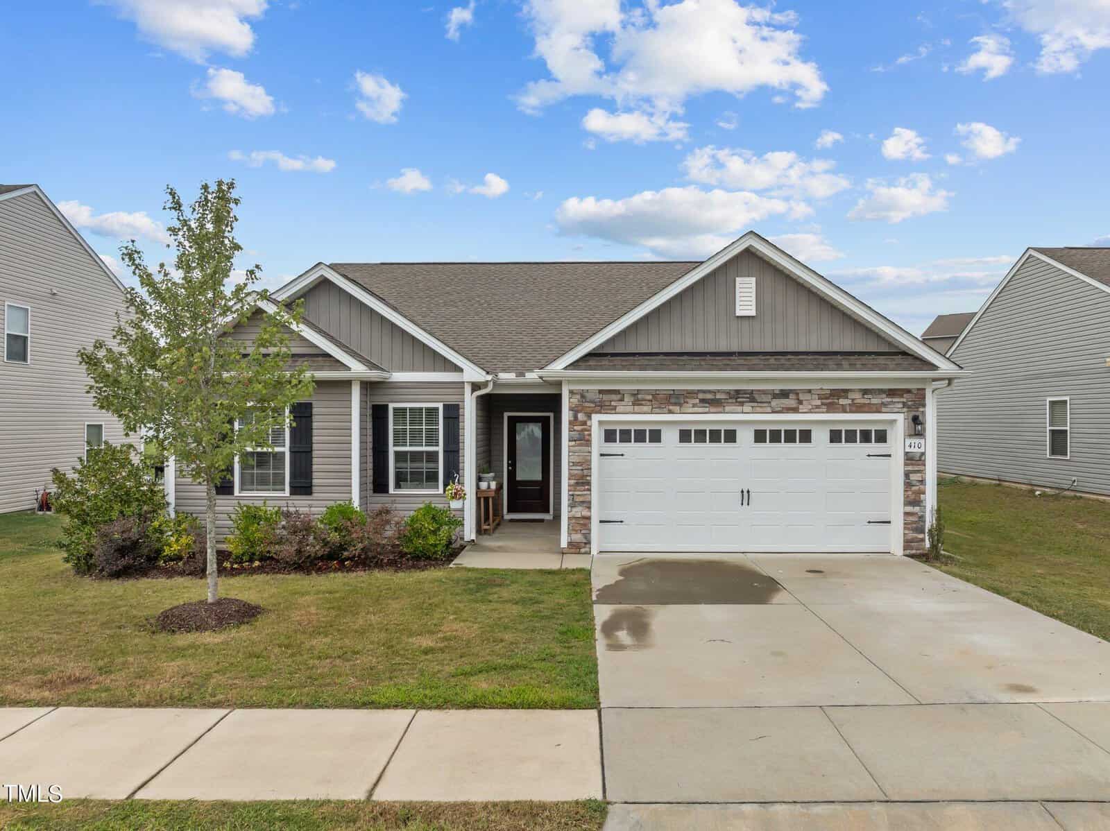 A single-story suburban house with gray siding, stone accents, and a two-car white garage. A small covered porch and a young tree are in the front yard, and the sky is partly cloudy.