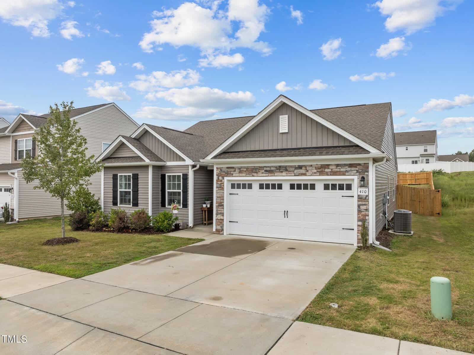 Single-story house with gray siding and stone accents, white trim, a double garage with white doors, small front yard with a tree, concrete driveway, and a wooden fence along the right side. Blue sky with scattered clouds above.