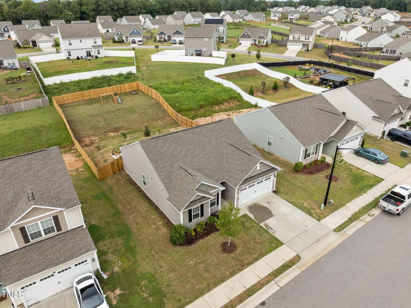 Aerial view of a suburban neighborhood featuring single-story houses with garages, neatly mowed lawns, driveways, and fenced backyards. Several houses are visible, arranged along a paved street.