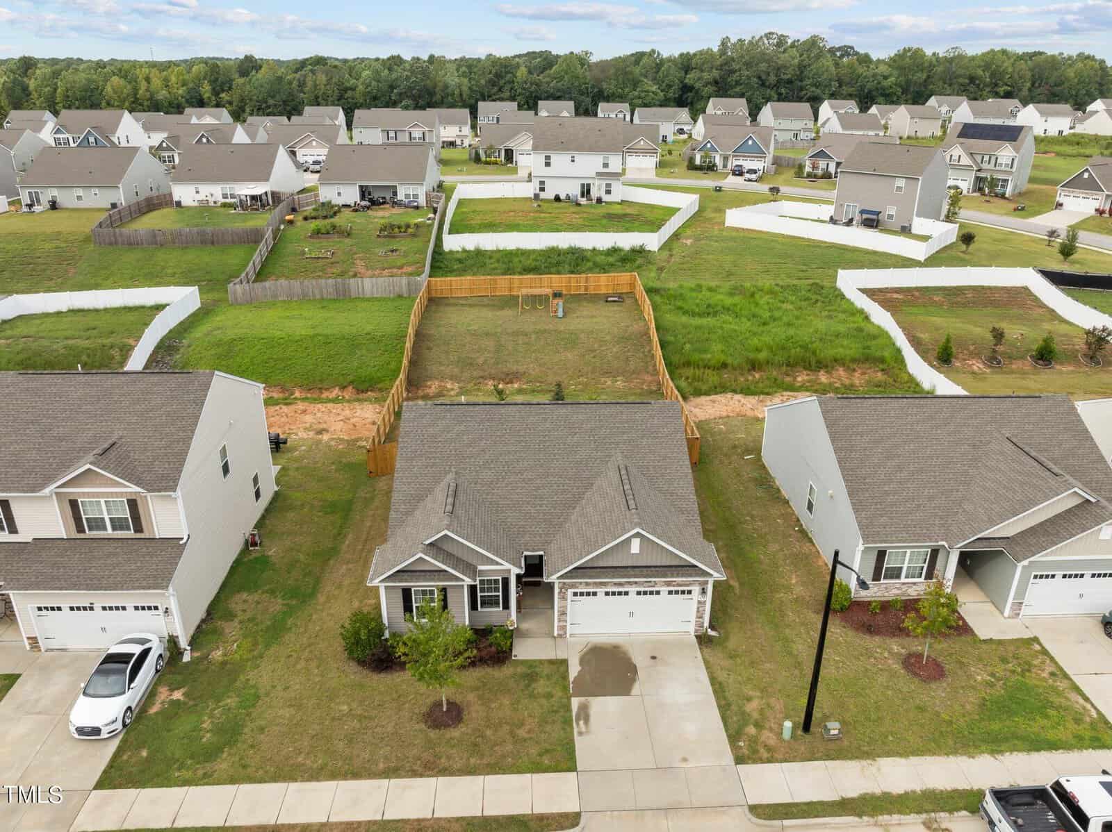 Aerial view of a suburban neighborhood with single-family homes; the central house has a fenced backyard, driveway, and gray roof. Similar houses with yards and driveways surround it, with green grass and trees in the background.