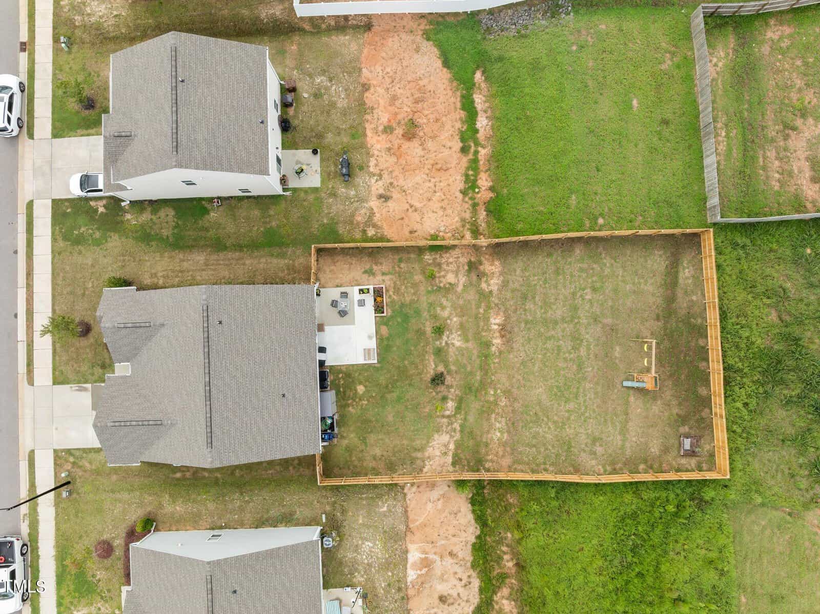 Aerial view of two houses with fenced and unfenced yards; one yard has a patio set and play equipment inside a wooden fence, while the other yard is open with visible patches of grass and dirt.
