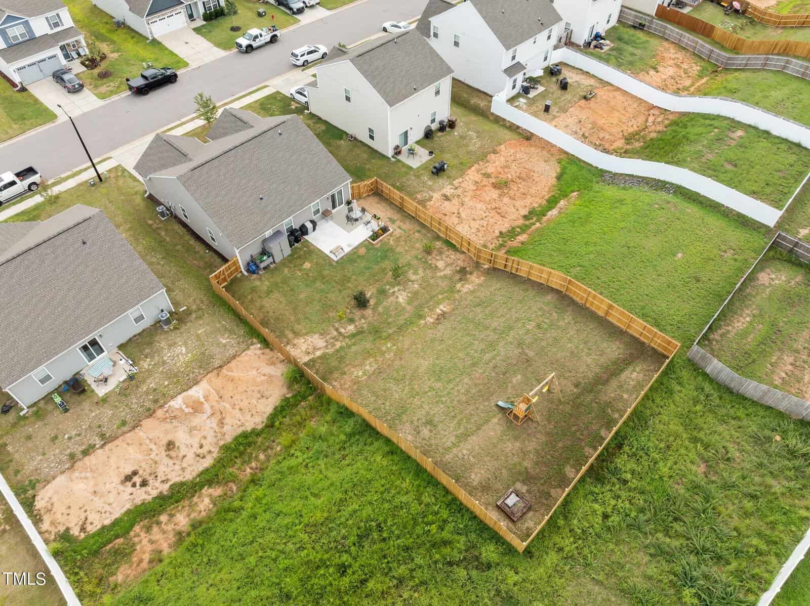 Aerial view of a fenced backyard with a small swing set, patio, and patchy grass. Surrounding houses have similar yards, some with bare soil areas. Residential neighborhood with detached single-family homes.