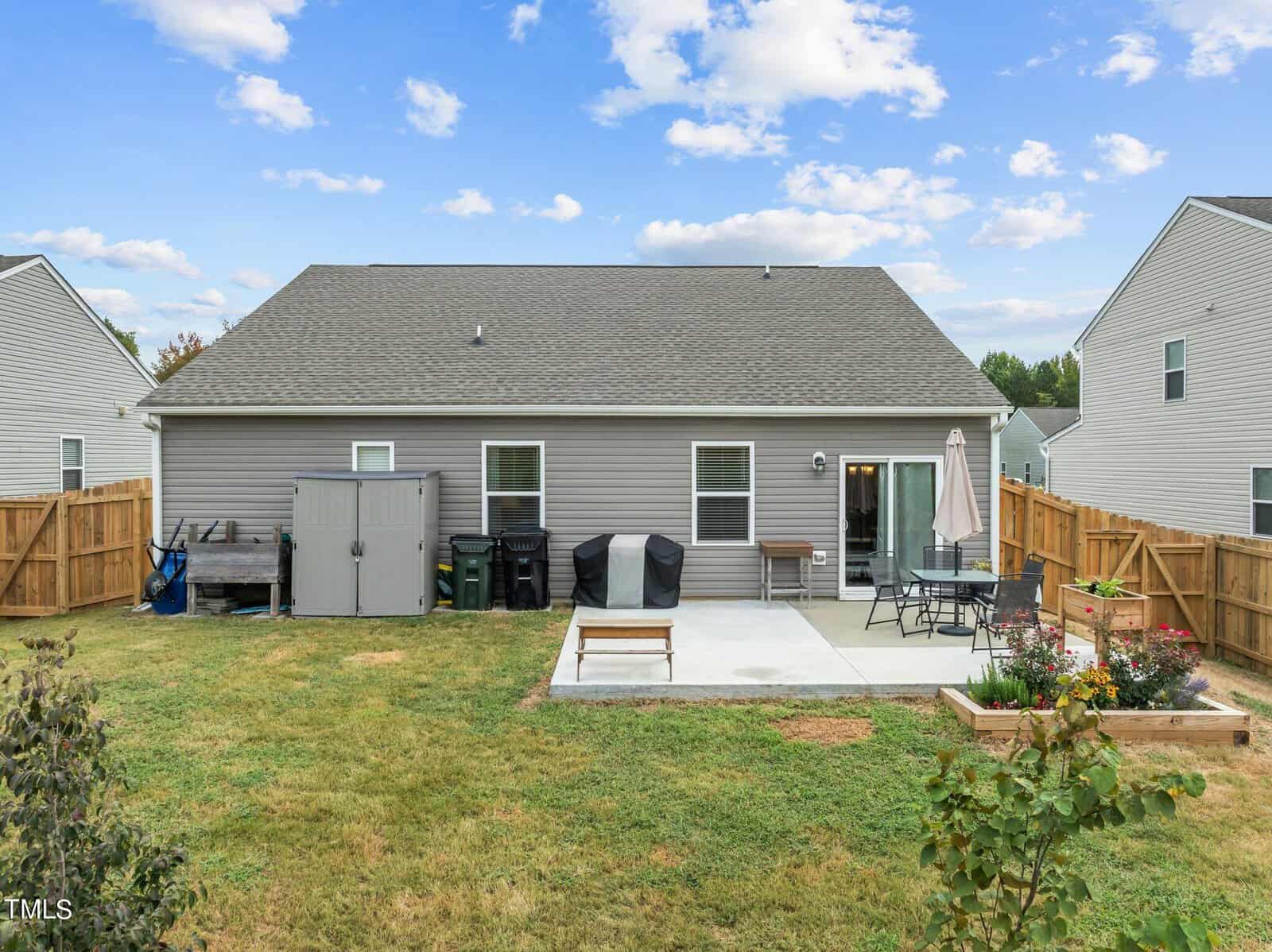 A fenced backyard with a gray single-story house, a concrete patio with two grills, a storage shed, patio furniture, a raised garden bed, grassy lawn, and partly cloudy sky.