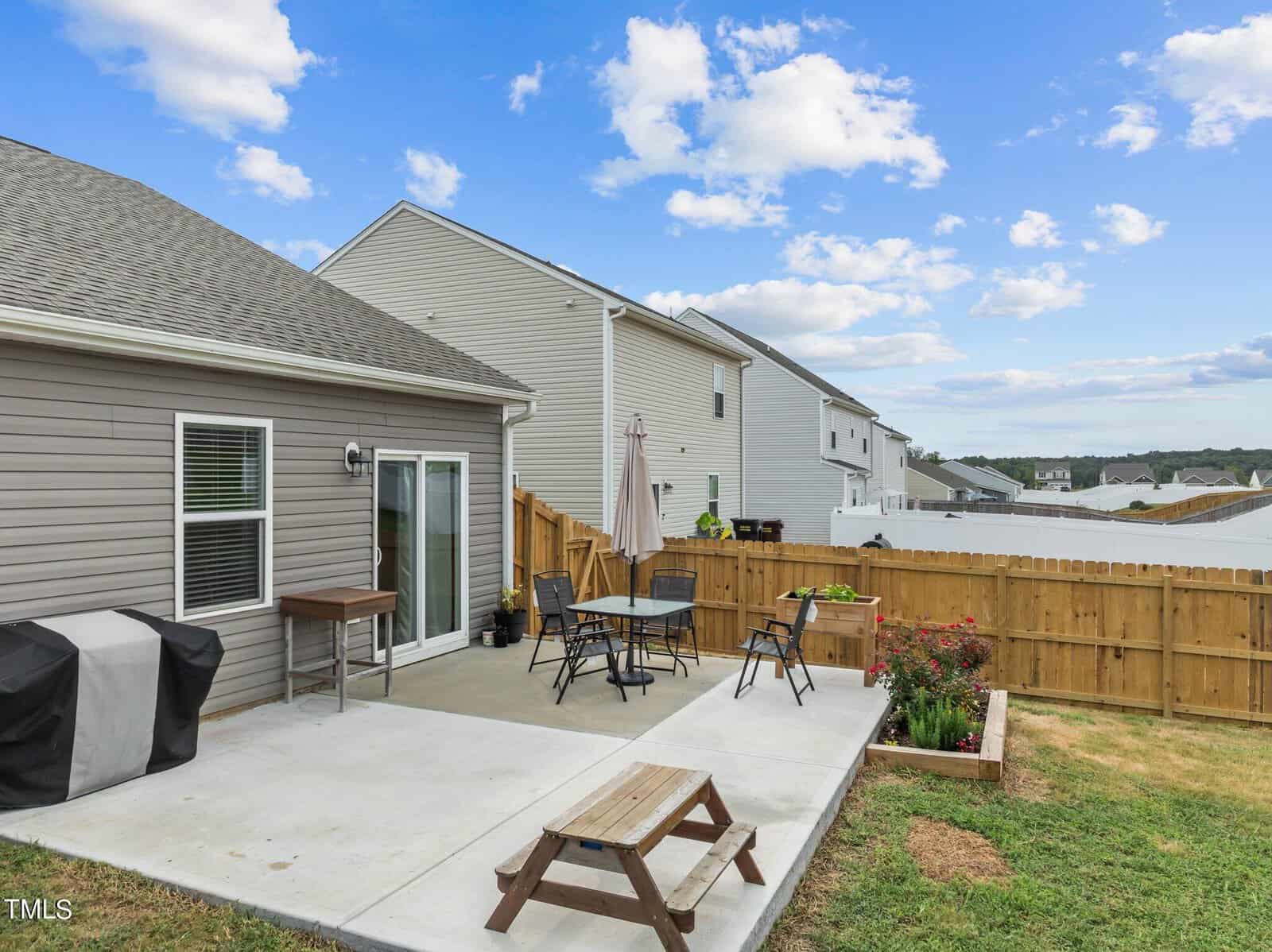 A fenced backyard with a concrete patio featuring a grill, a dining table with chairs, an umbrella, potted plants, and a small wooden picnic table. Two houses and a partly cloudy sky are visible in the background.