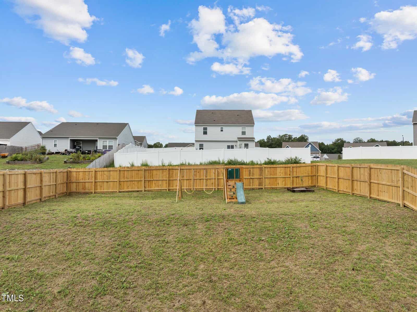 A fenced backyard with a small playset including a slide and swings, surrounded by grass. Neighboring houses and a blue sky with scattered clouds are visible in the background.