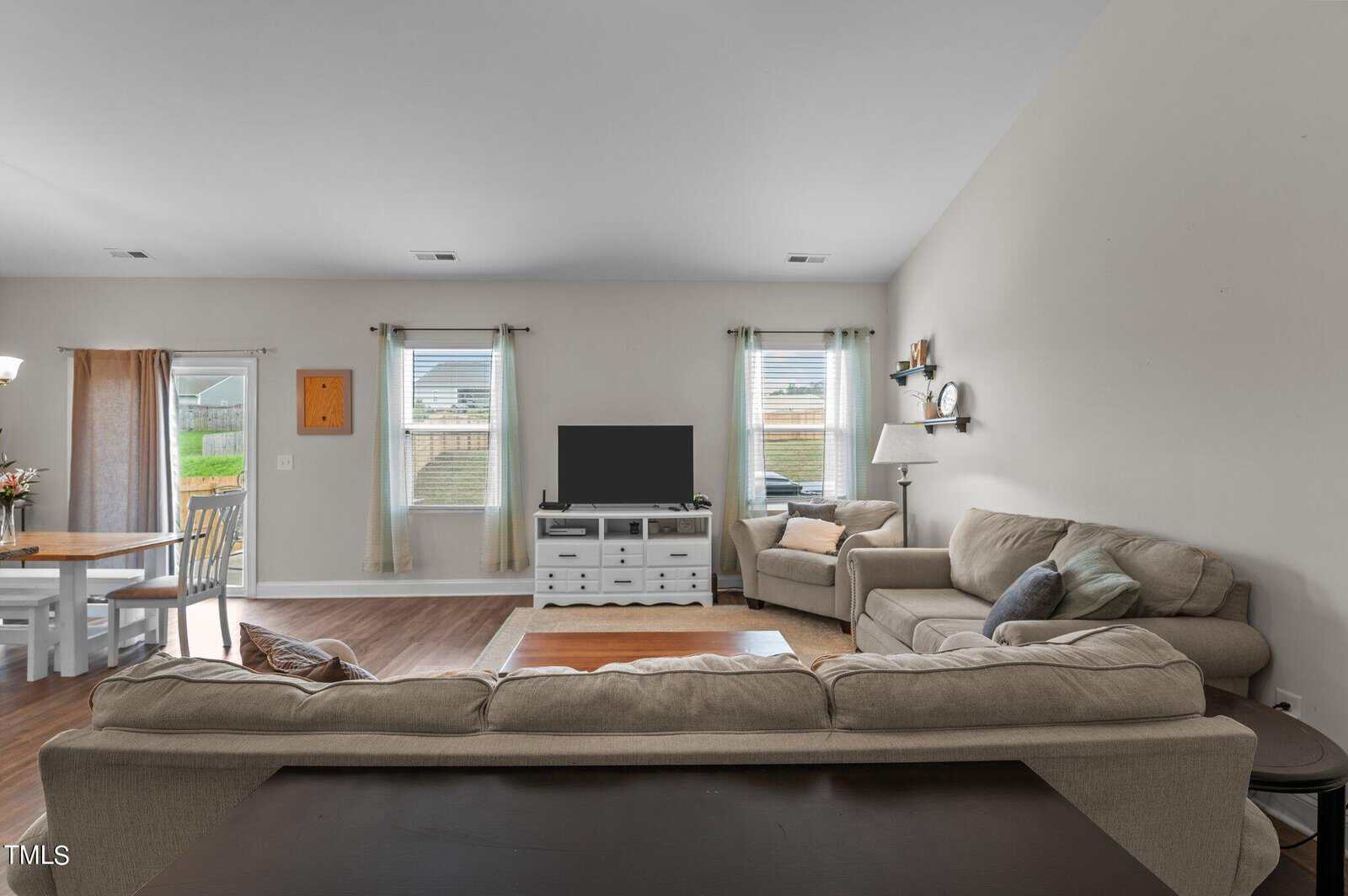 A living room with beige sofas, a TV on a white stand, a small rug, and windows with light curtains. A dining area with a table and chairs is visible to the left, and the walls are painted light gray.