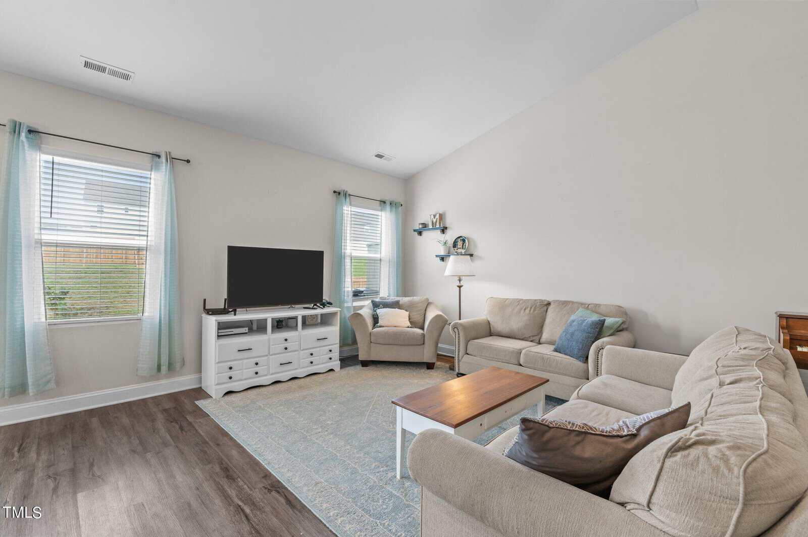 Bright living room with two beige sofas, an armchair, a coffee table, a white TV stand with a TV, blue patterned rug, wood floors, and two windows with light blue curtains.