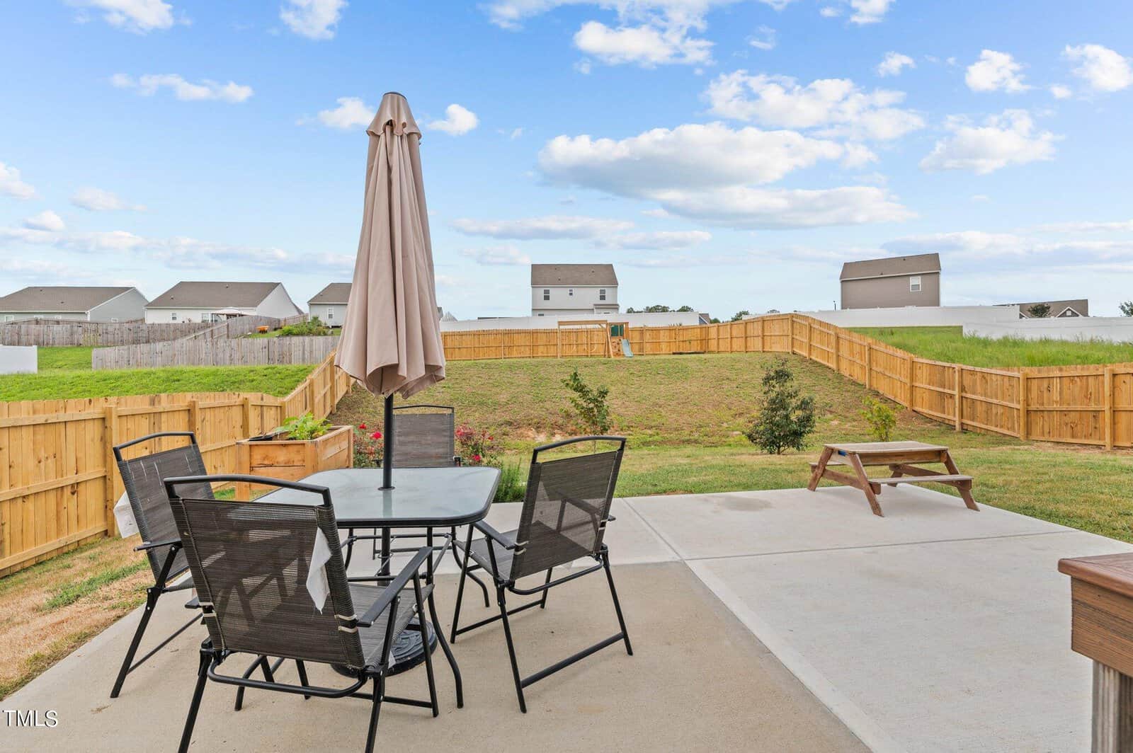 A backyard with a patio set and umbrella, a wooden picnic table, a lawn, flowers, and a wooden privacy fence. Houses and a blue sky with clouds are in the background.