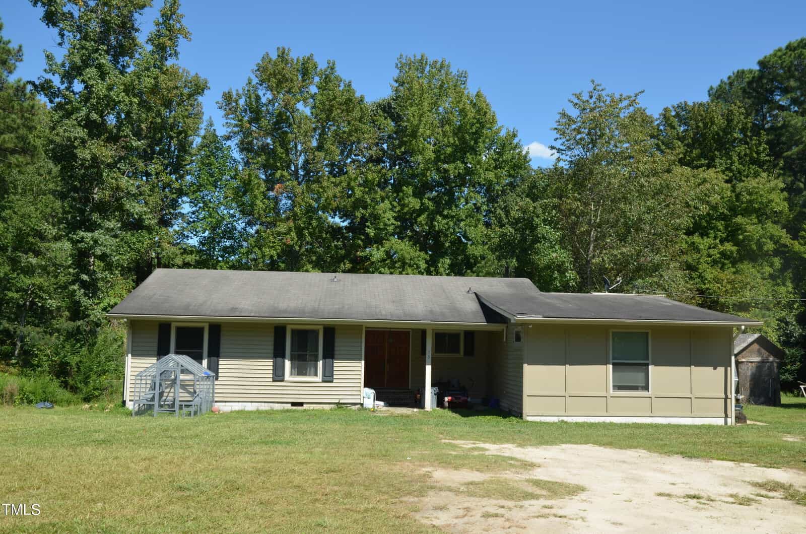 Single-story house with beige siding and a dark roof, set in front of a wooded area. There is a small green greenhouse on the lawn and a dirt driveway in front. The house has four visible windows and a covered entryway.