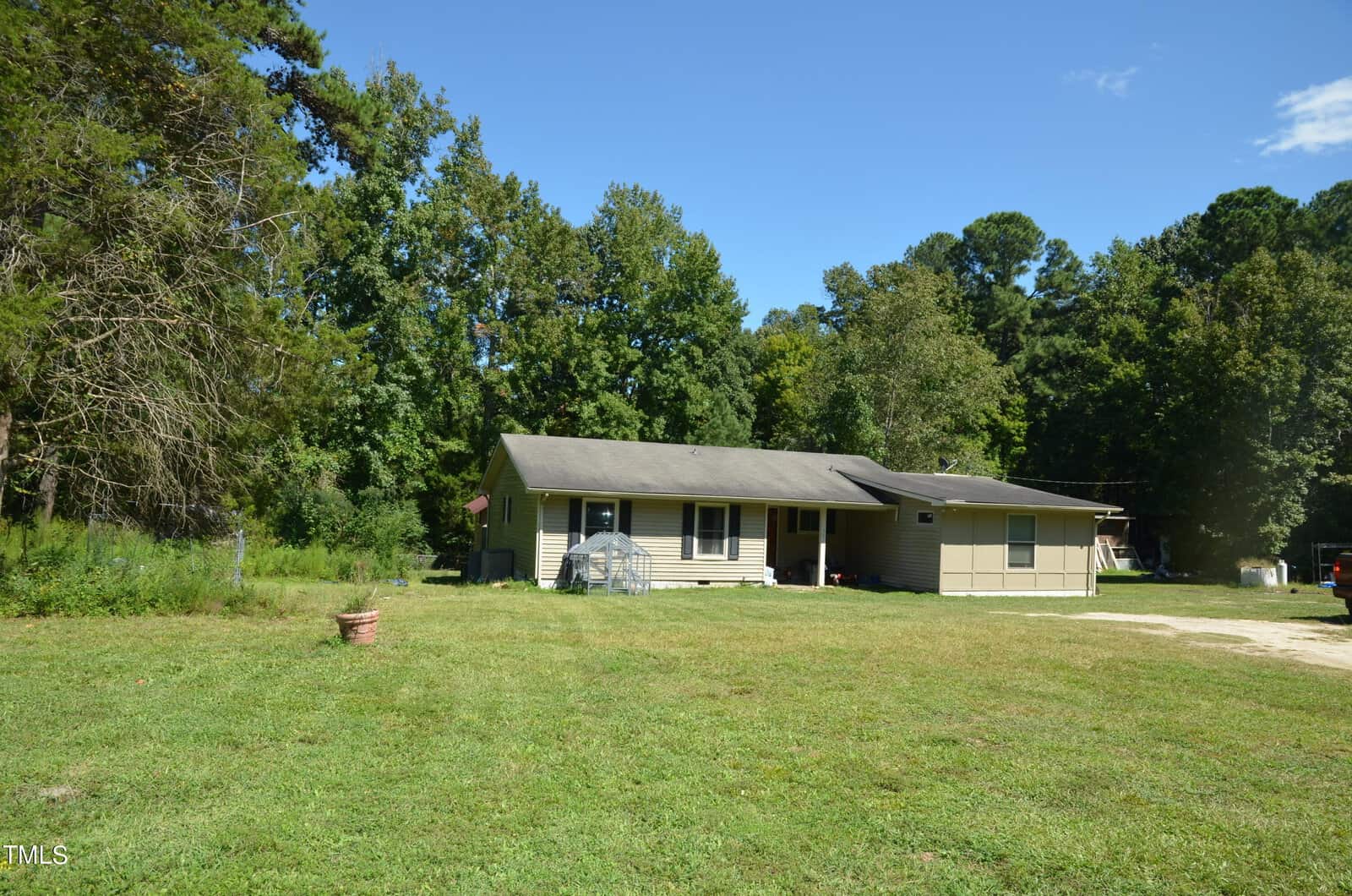 Single-story house with a covered front porch, tan and olive siding, set on a large grassy yard bordered by tall trees under a partly cloudy sky. A potted plant and a small garden structure are visible in the yard.